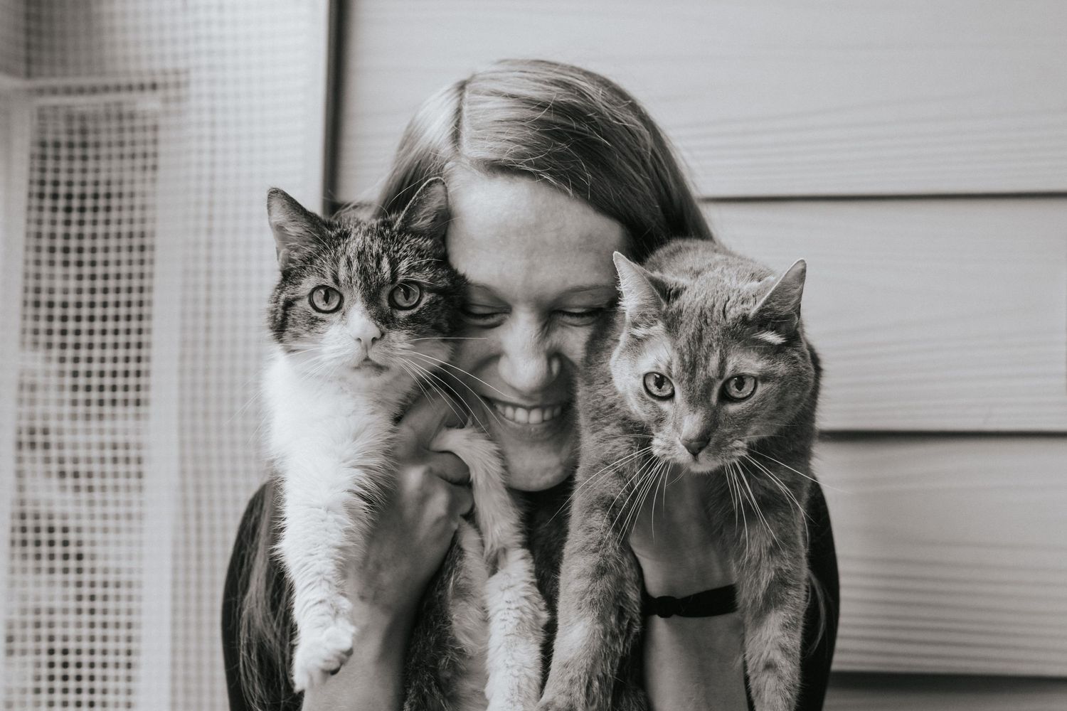 A black and white photo of a smiling woman holding two cats close to her face.