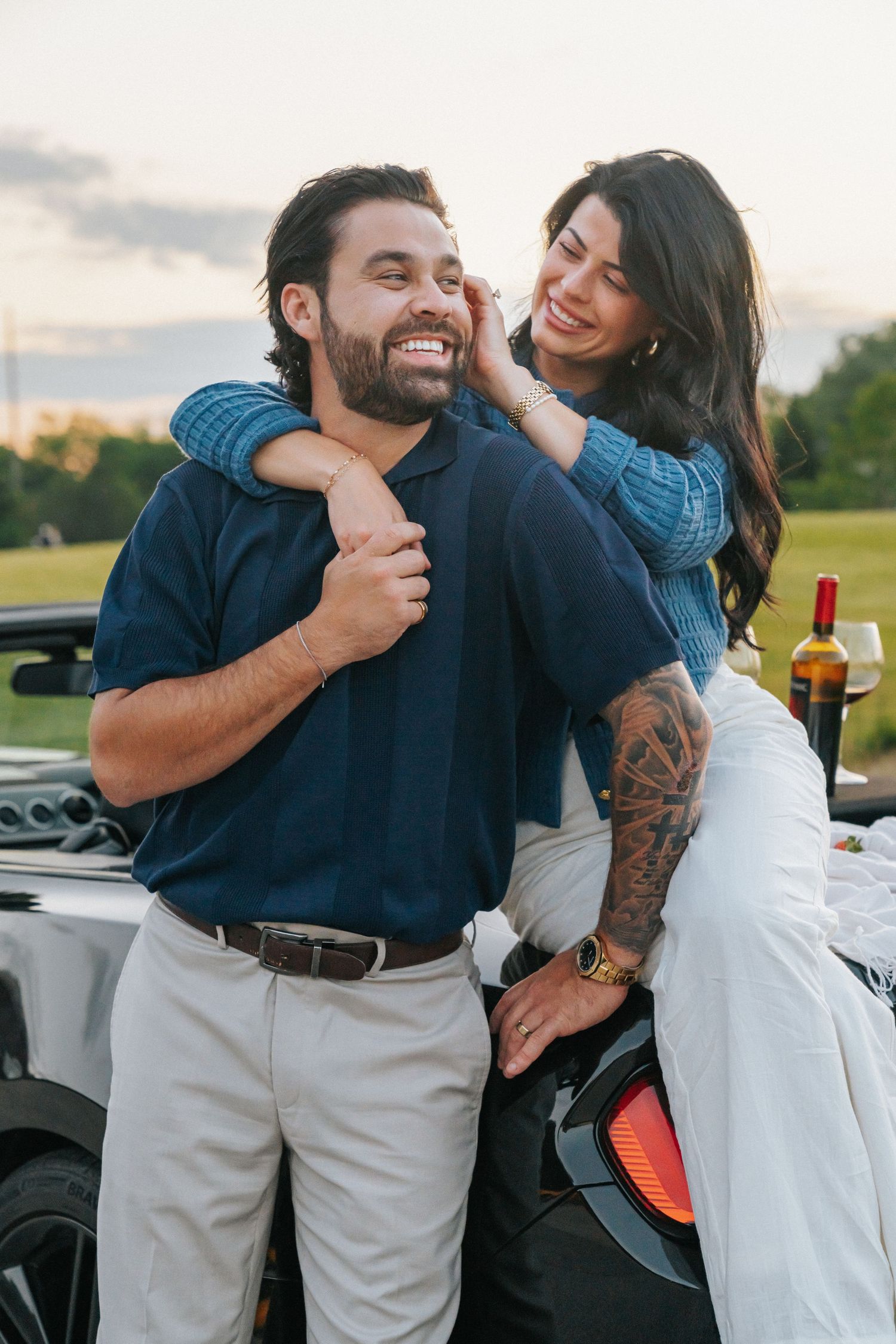 A smiling couple embraces beside a convertible car at sunset, woman with tattoos hugging man from behind.