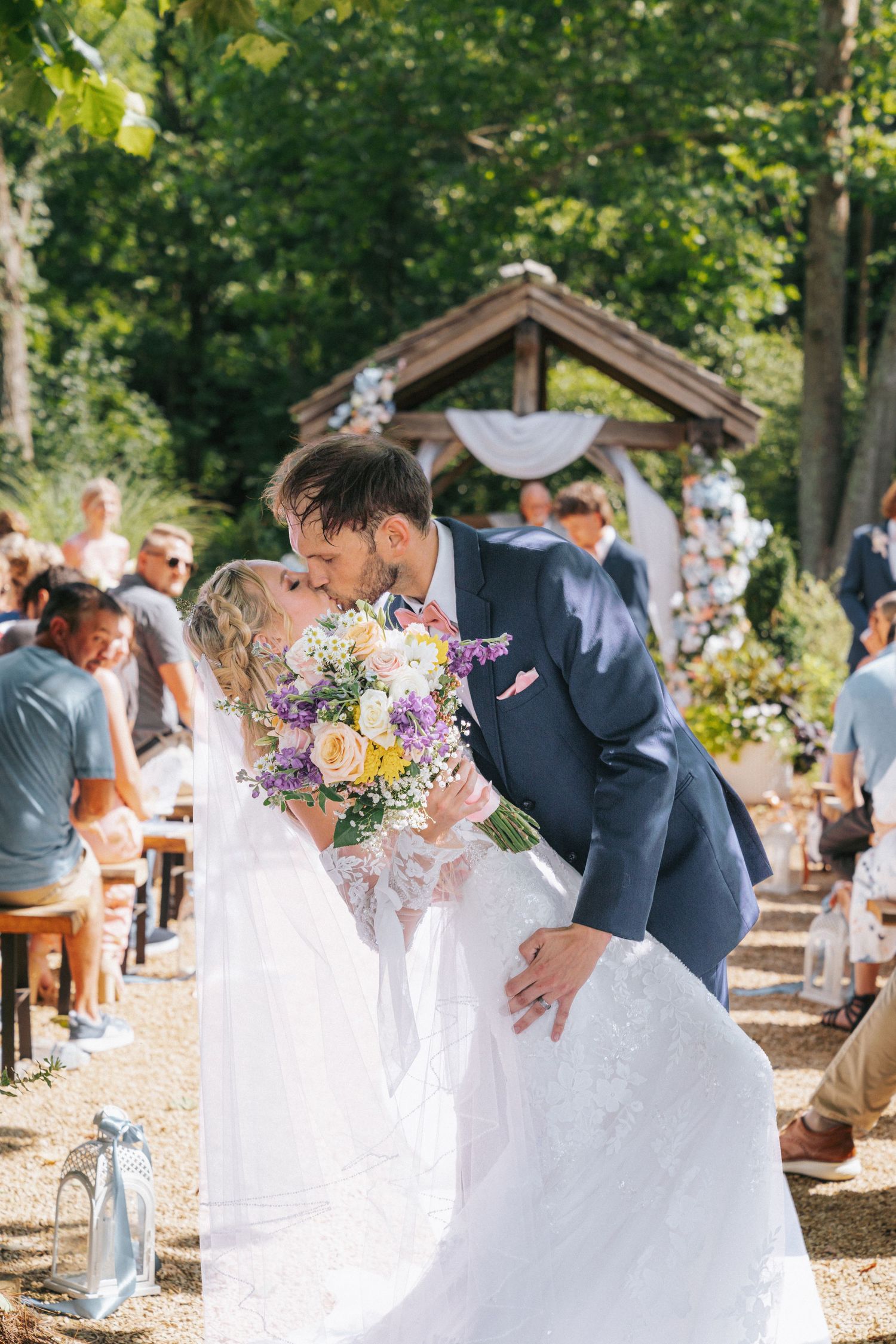 Bride and groom share a romantic kiss at an outdoor wedding ceremony surrounded by guests and lush green trees.