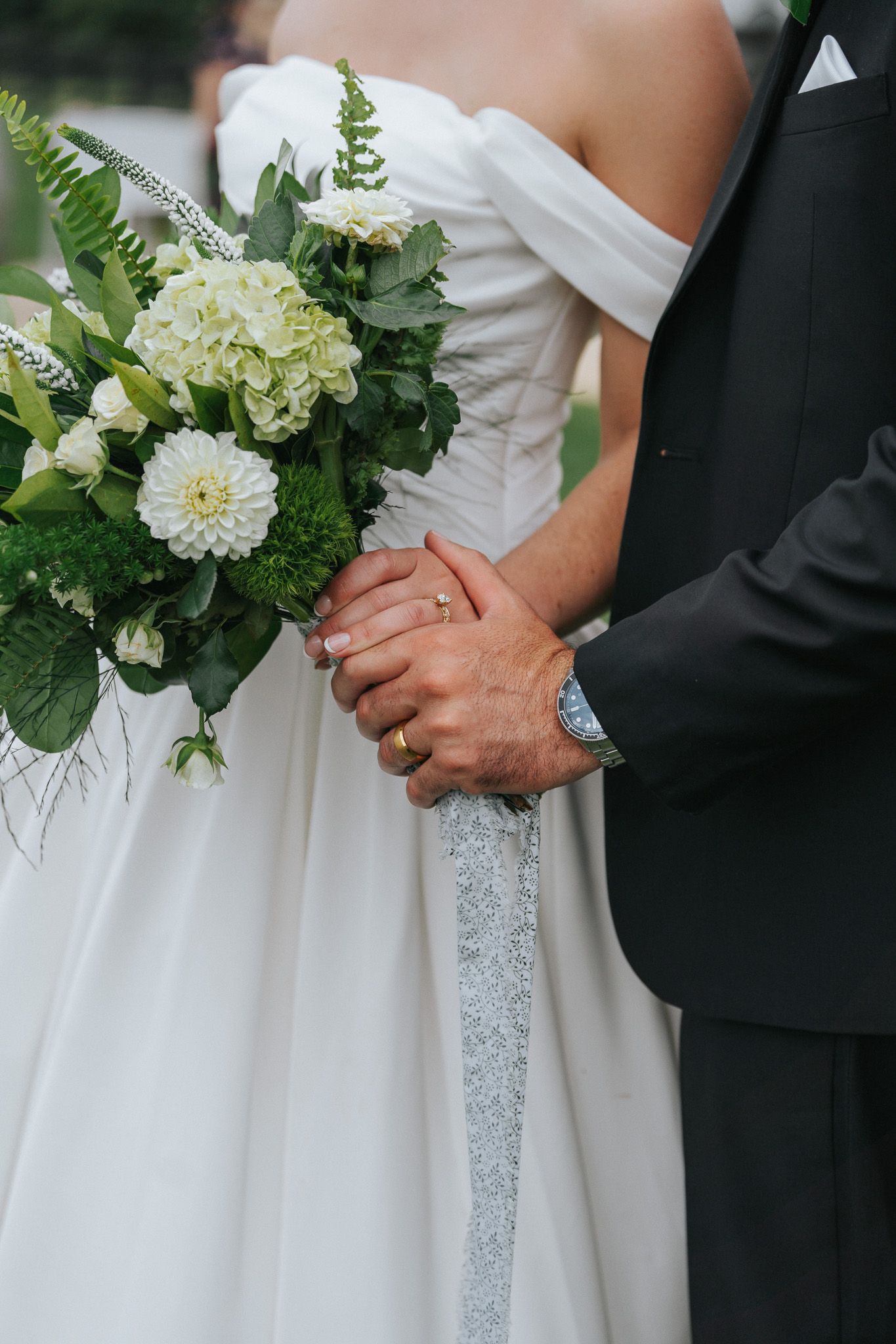 Bride and groom holding hands at wedding, bride holding white hydrangea and dahlia bouquet with greenery, wearing wedding rings.
