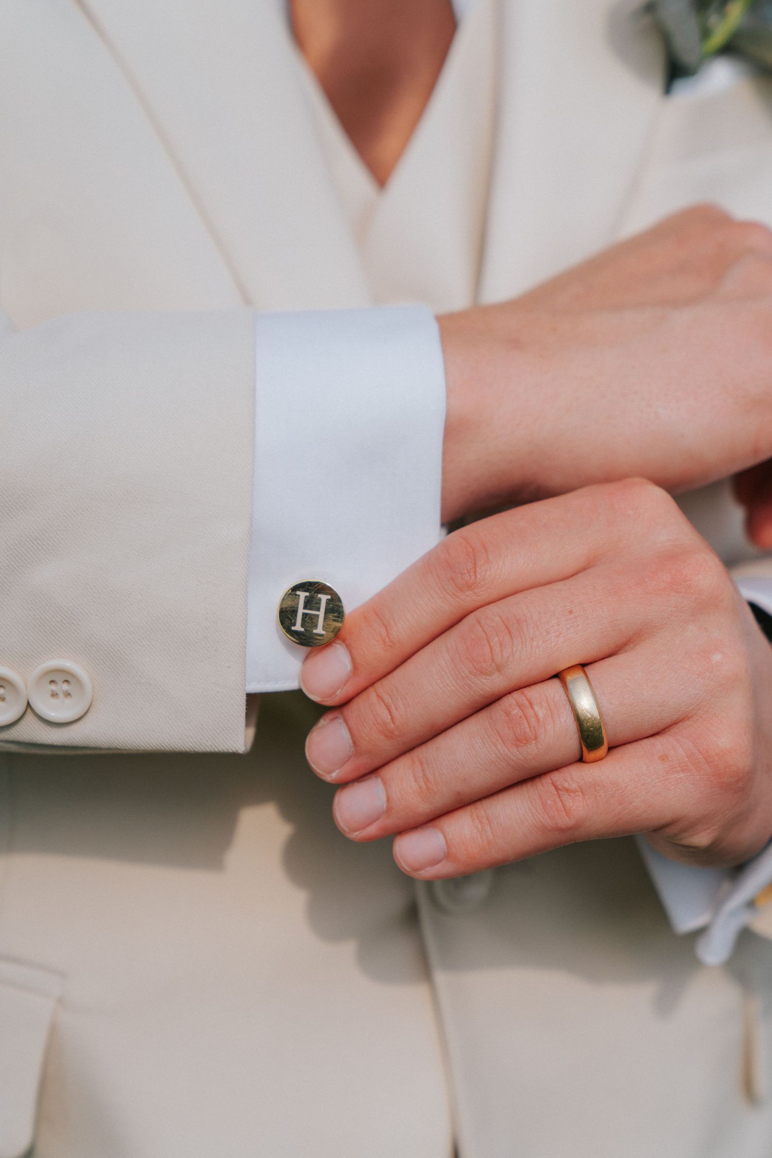 Close-up of groom adjusting gold H monogram cufflink, wearing wedding band and cream suit.