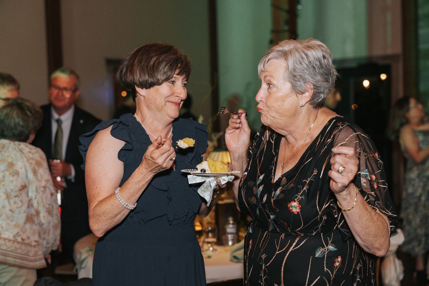 Two elderly women in elegant attire chat and share dessert at a formal event or wedding reception.