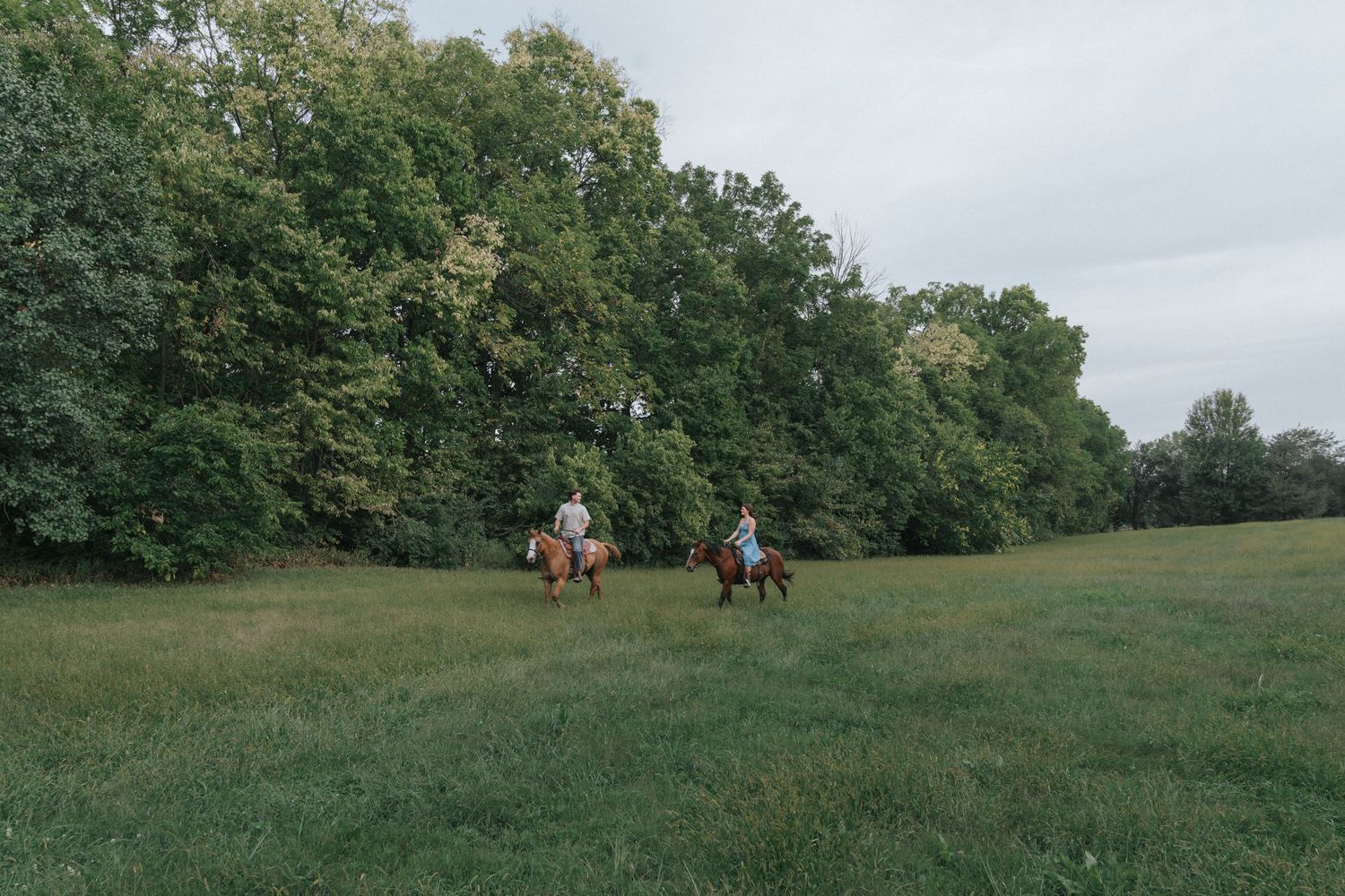 Two riders on horseback cross a wide green meadow bordered by dense trees on a cloudy day.