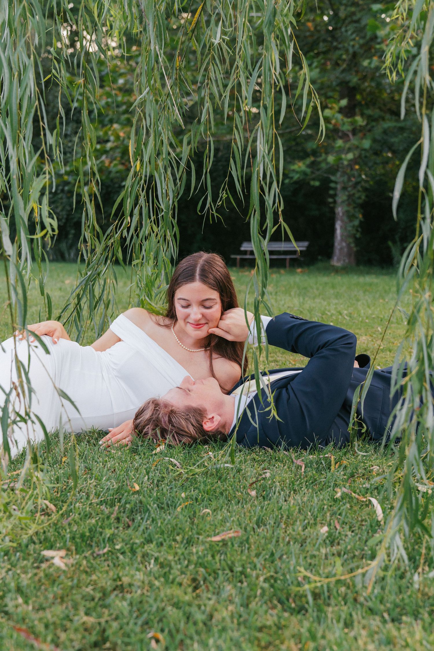 Couple in wedding attire lying on green grass under a weeping willow tree in a romantic outdoor setting.