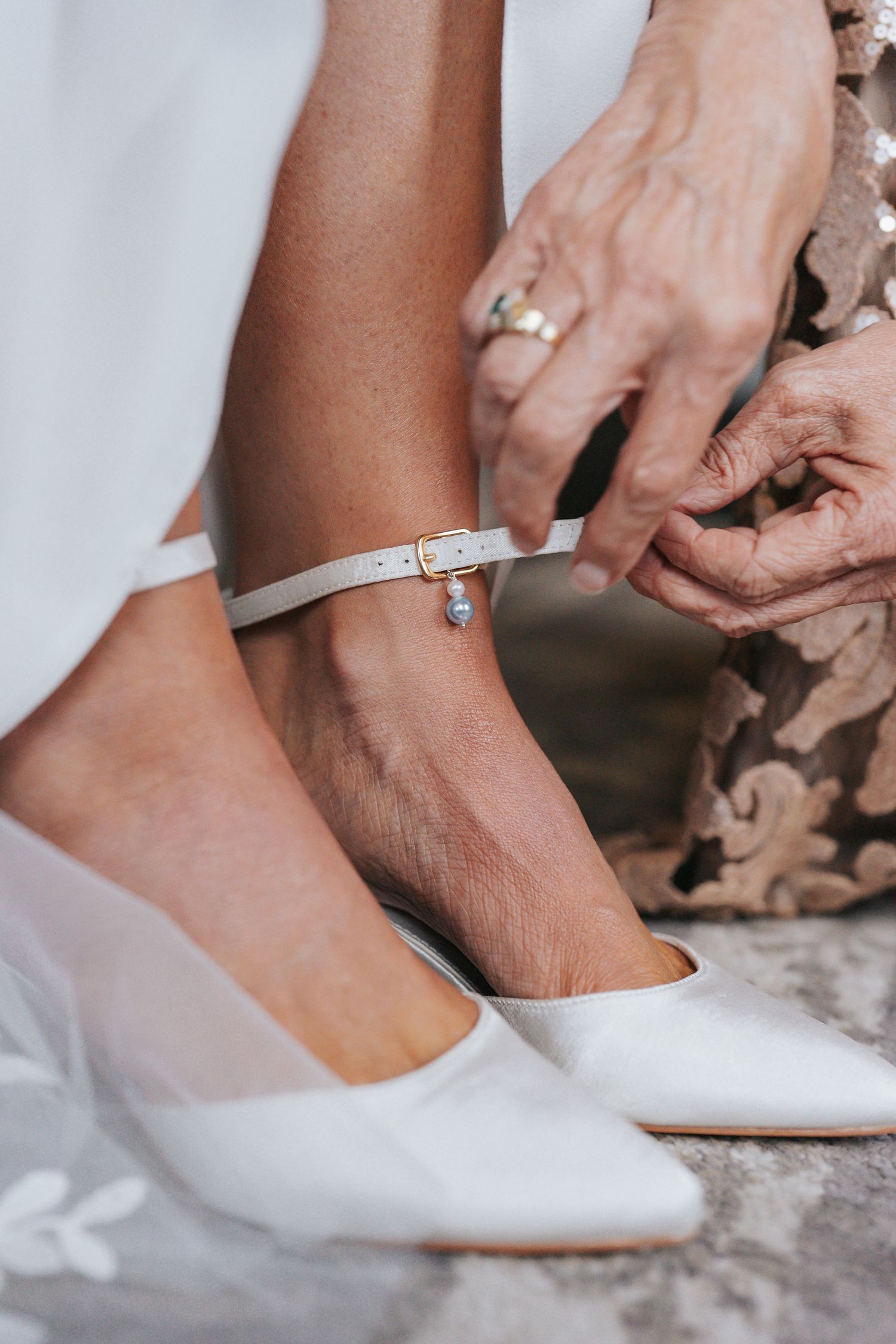 Helper fastening white ankle strap bridal heel with pearl charm on bride.