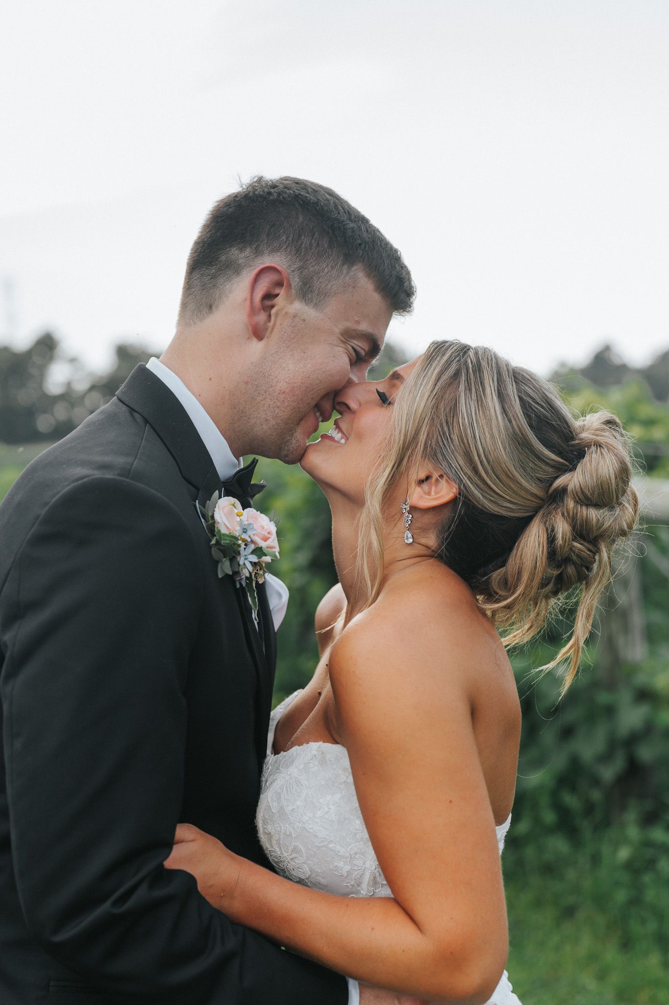 A bride and groom share a romantic almost-kiss moment outdoors, dressed in wedding attire with a floral boutonniere.