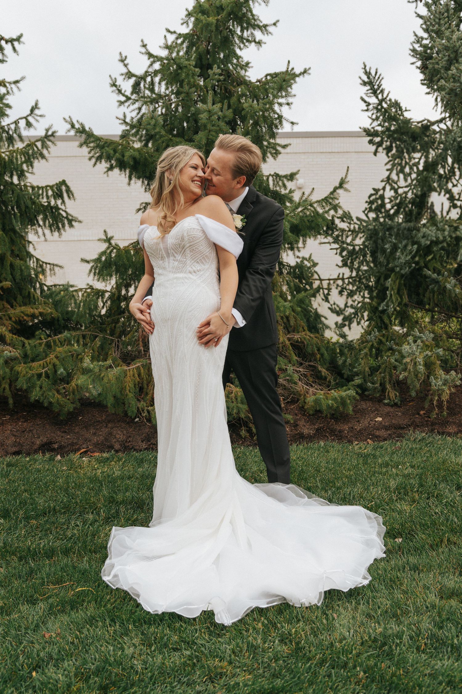 Bride and groom share a romantic embrace outdoors, with the bride in an elegant off-shoulder white gown with a flowing train.