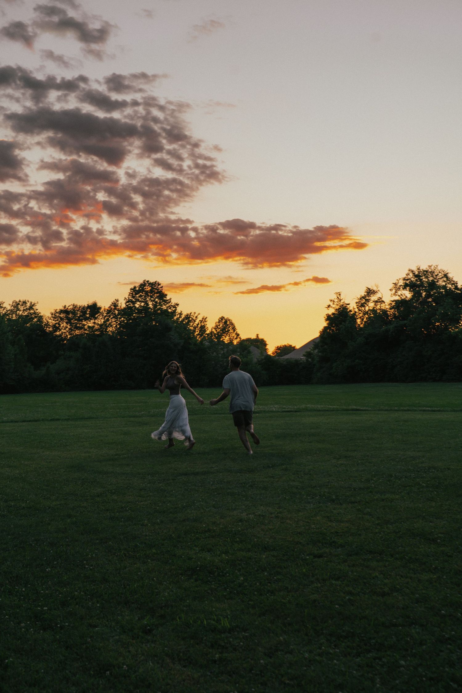 Couple holding hands running across a green field at sunset with colorful clouds.