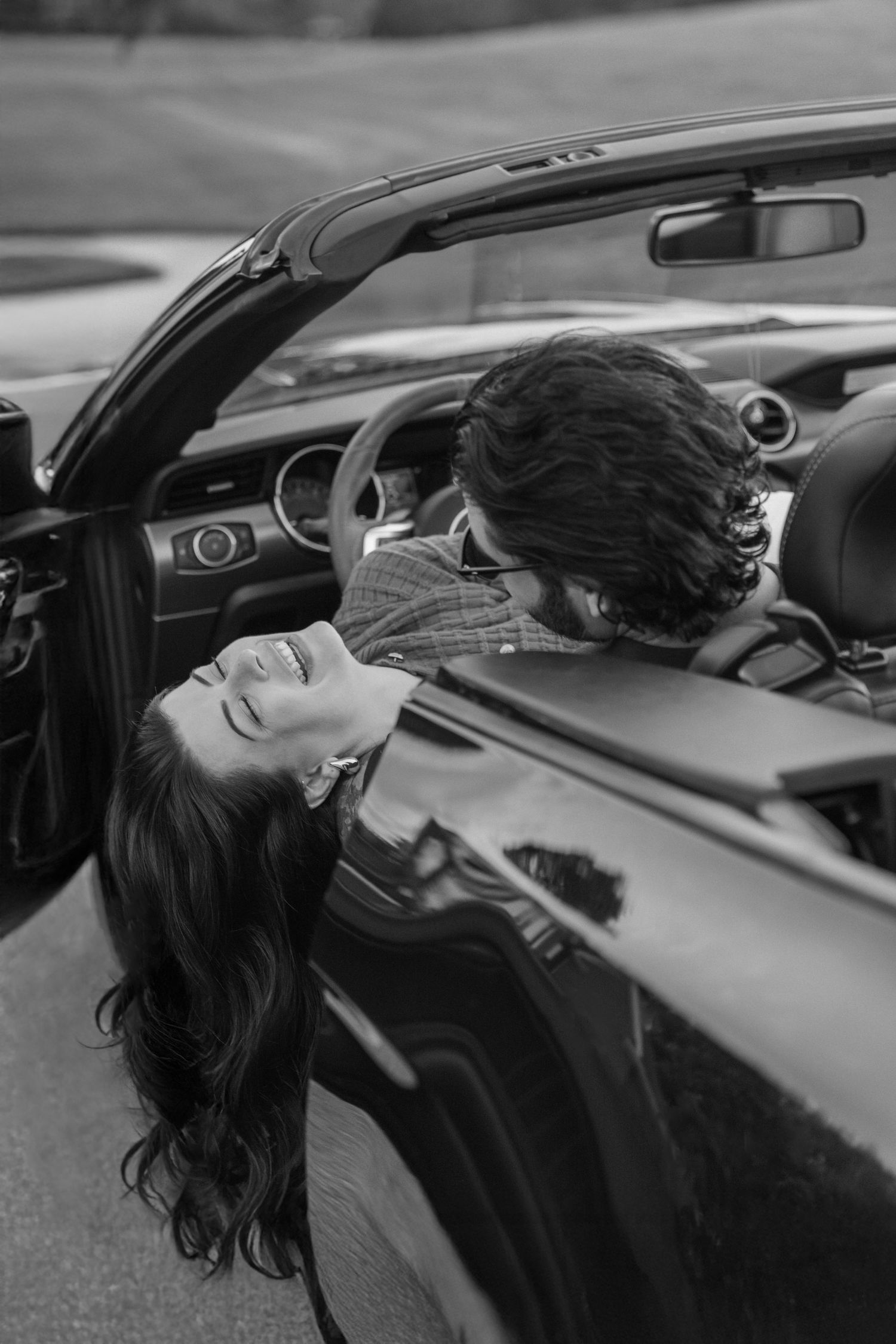 Black and white photo of a couple in a convertible car, woman leaning back laughing joyfully.