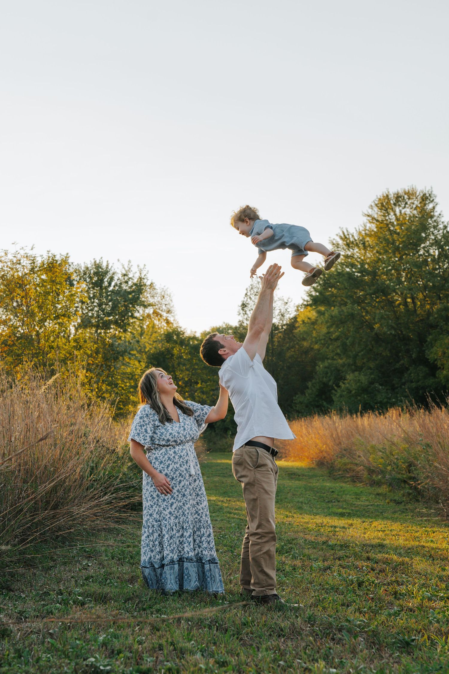 Father joyfully tossing toddler in the air while mother watches in a golden sunset meadow.