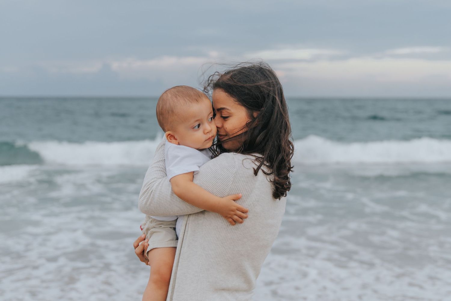 Loving mother holding and cuddling her baby boy on a cloudy beach with ocean waves behind them.