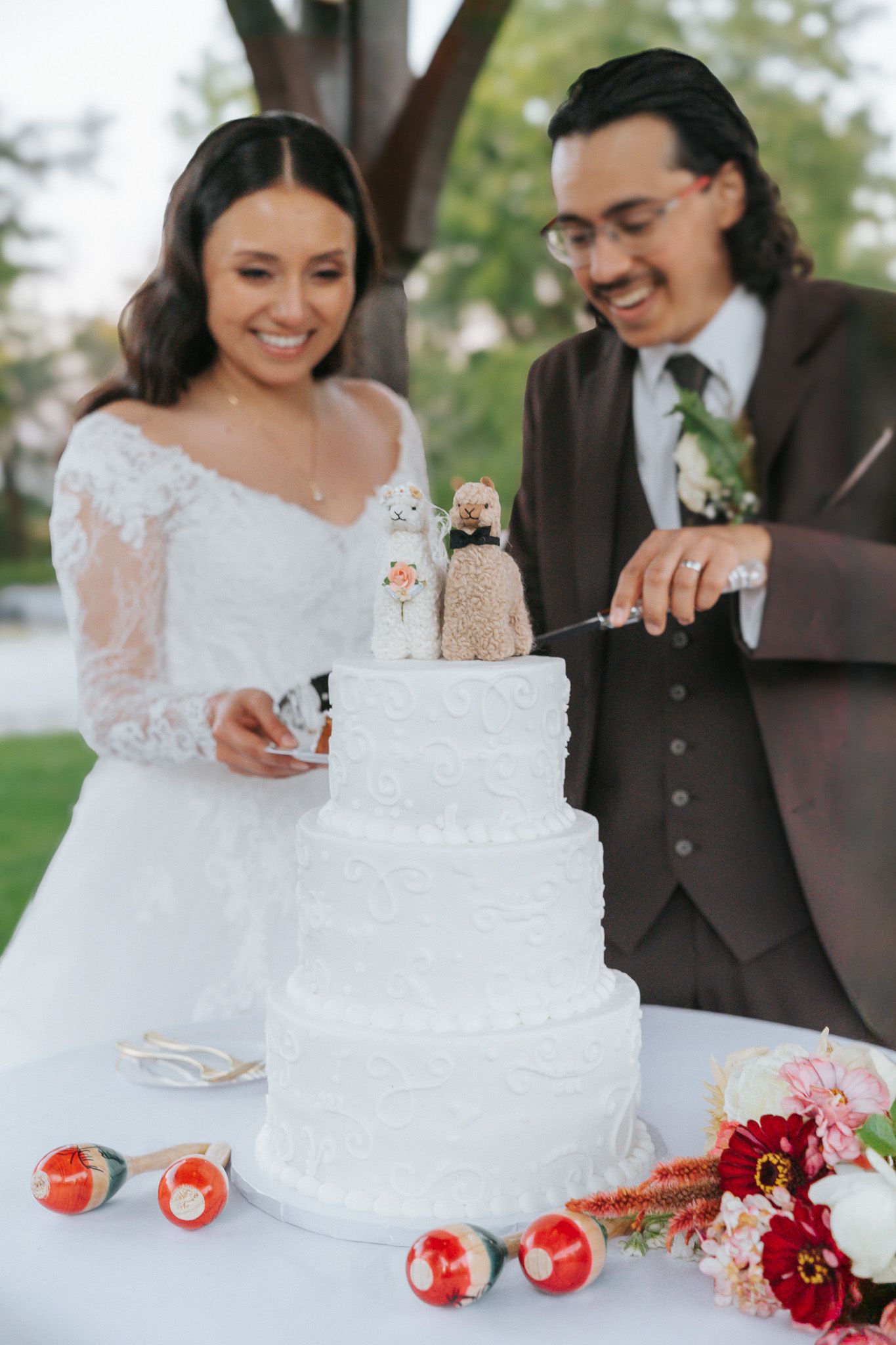 Smiling bride and groom cutting a three-tier white wedding cake topped with custom figurines at an outdoor reception.