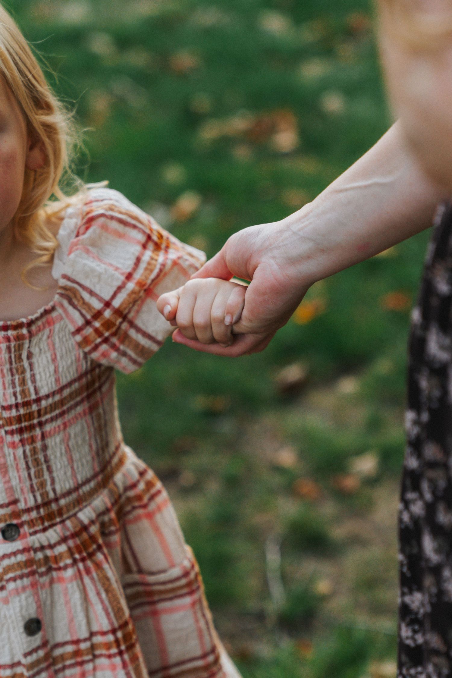 A mother tenderly holds her young daughter's hand outdoors, child wearing a plaid dress on green grass.