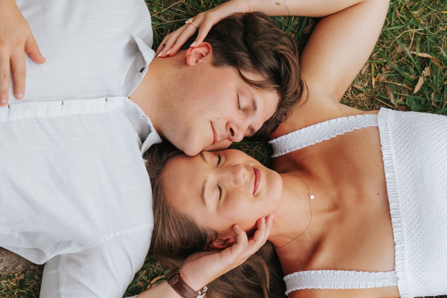Romantic couple lying on grass with eyes closed, sharing a tender moment outdoors in white summer outfits.