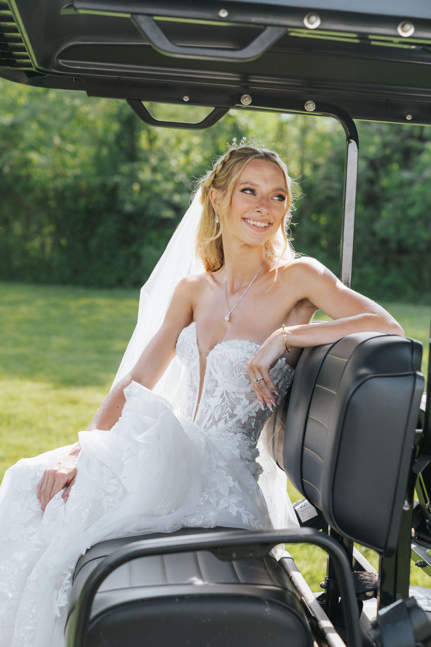 Smiling bride in white lace gown and veil sitting in a golf cart on a sunny green lawn.