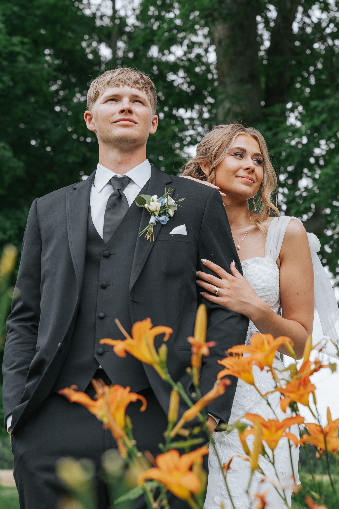Bride and groom posing outdoors near orange lilies, groom in black suit with boutonniere, bride in white dress smiling.