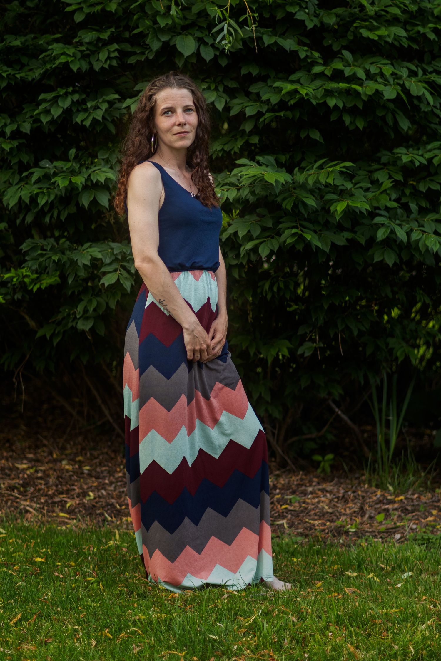 Woman in chevron maxi dress standing outdoors against green foliage backdrop.