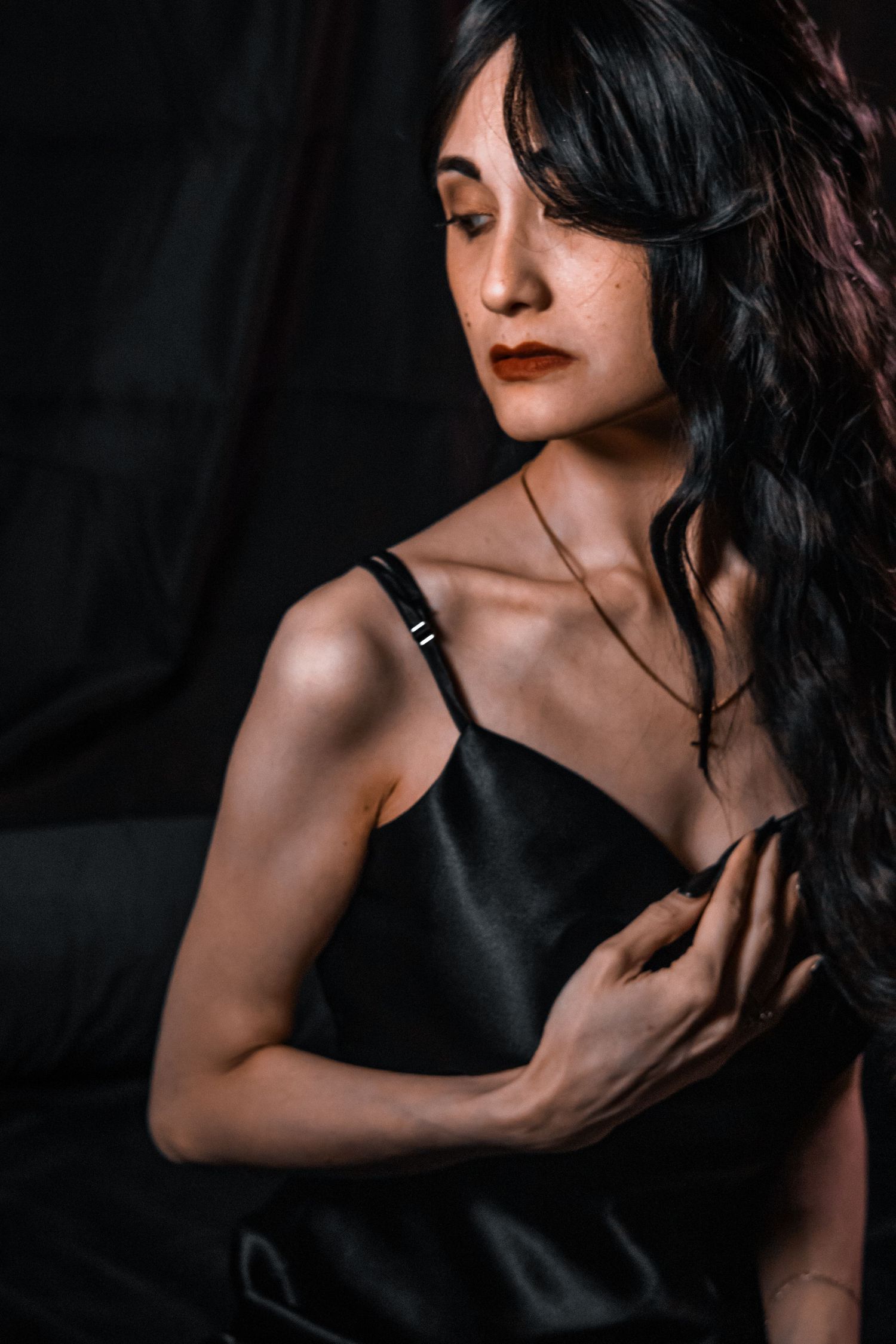 Woman in black satin slip dress with long dark wavy hair and red lips poses against dark background.