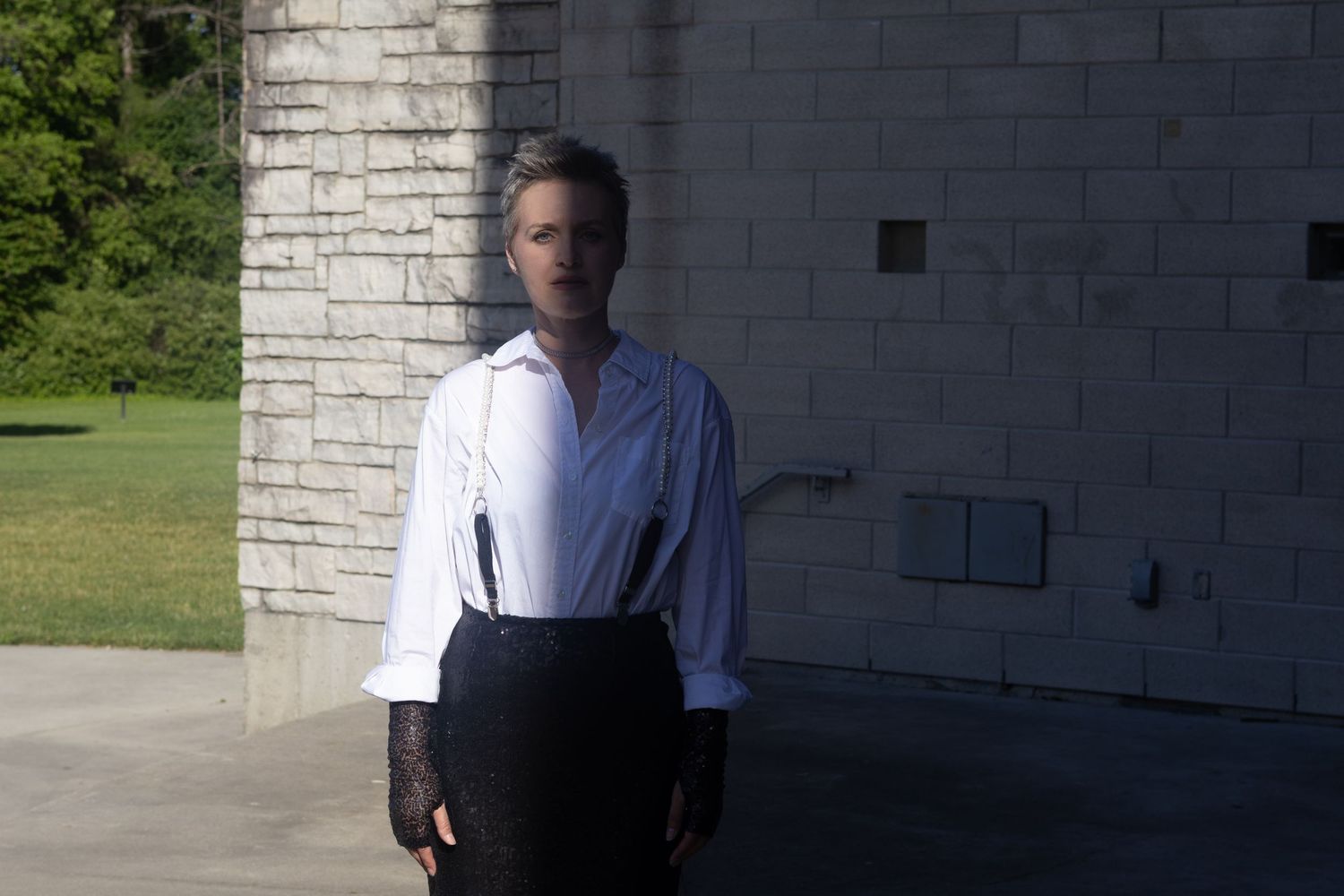 Person in white shirt and black skirt with suspenders stands outdoors near a stone building.