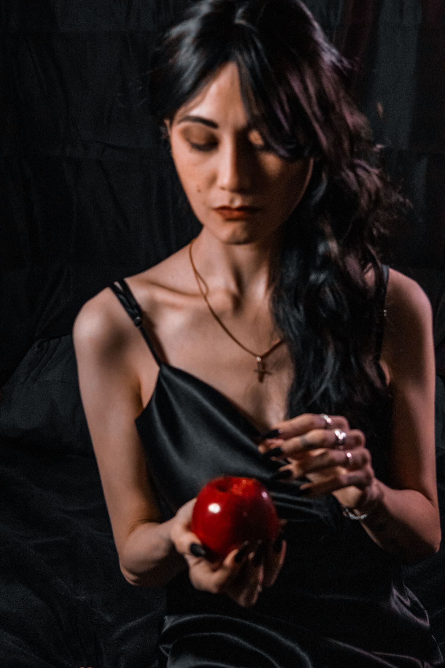 Young woman in black dress holding a red apple, wearing a cross necklace against dark background.