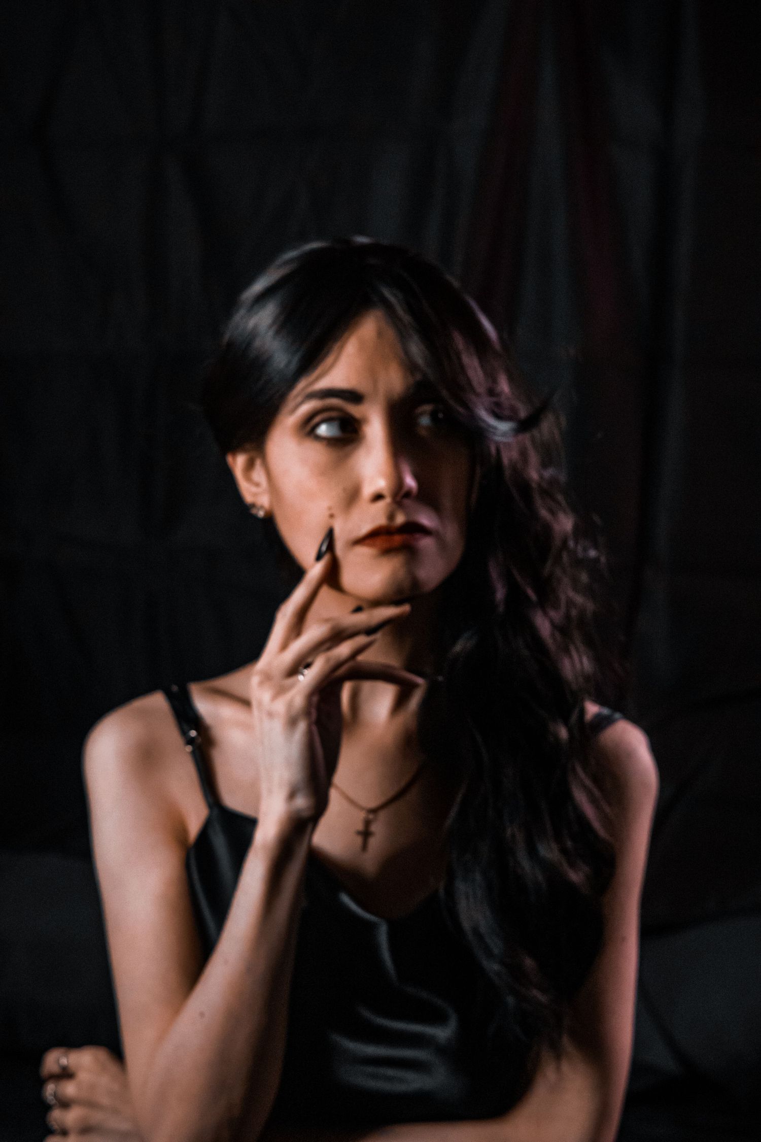 Woman in black outfit poses thoughtfully against dark backdrop, wearing cross necklace, with long dark wavy hair.