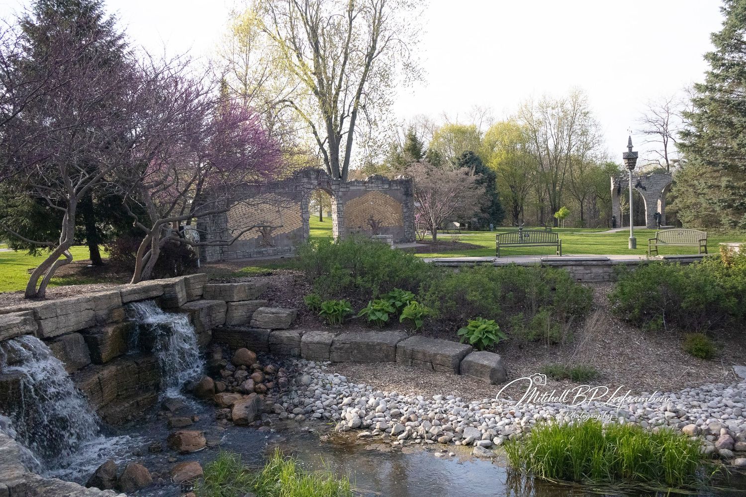 Scenic garden park with a small waterfall, stone walls, and blooming spring trees.