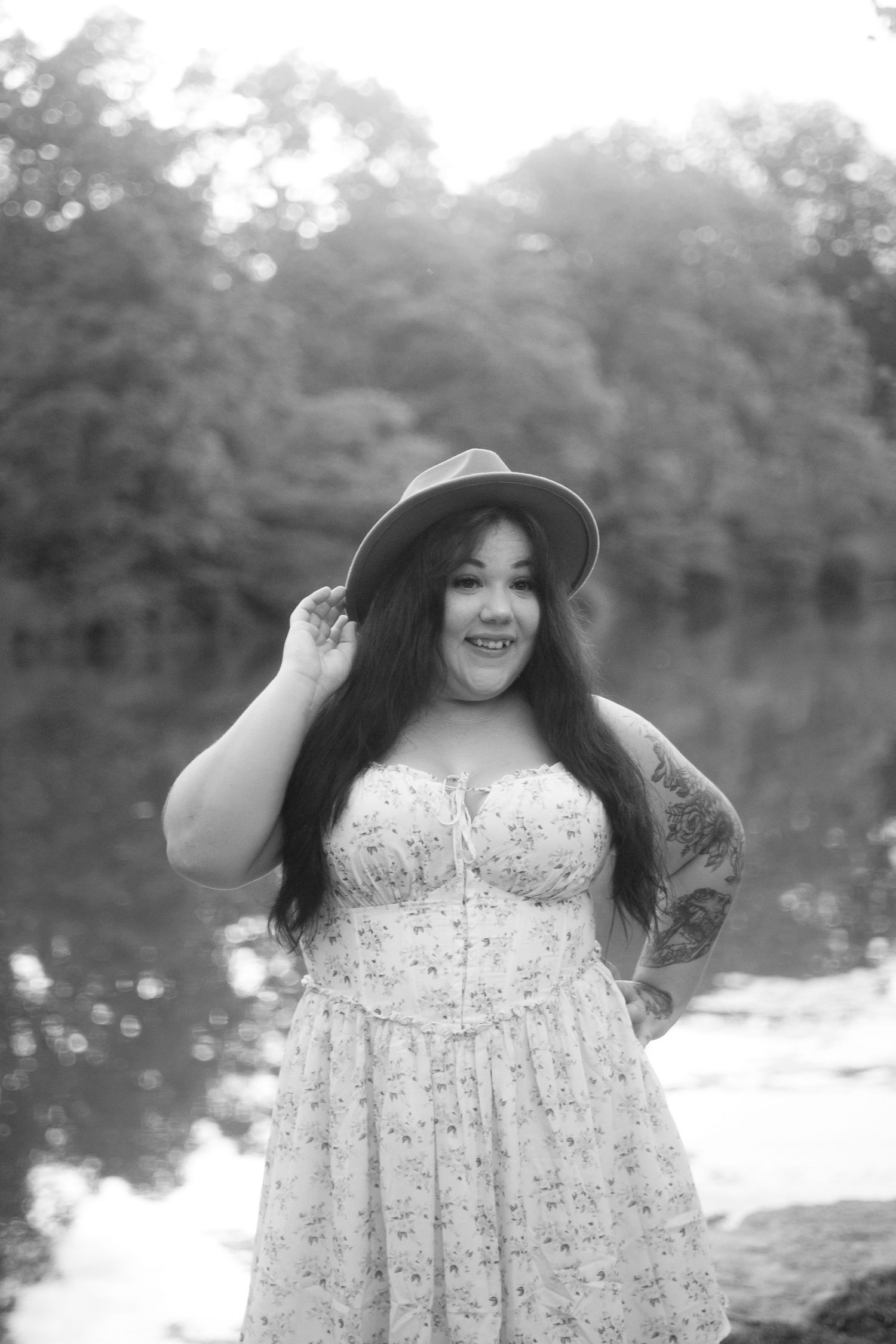 Black and white photo of a plus-size woman in floral dress and hat posing by a reflective lake with trees.