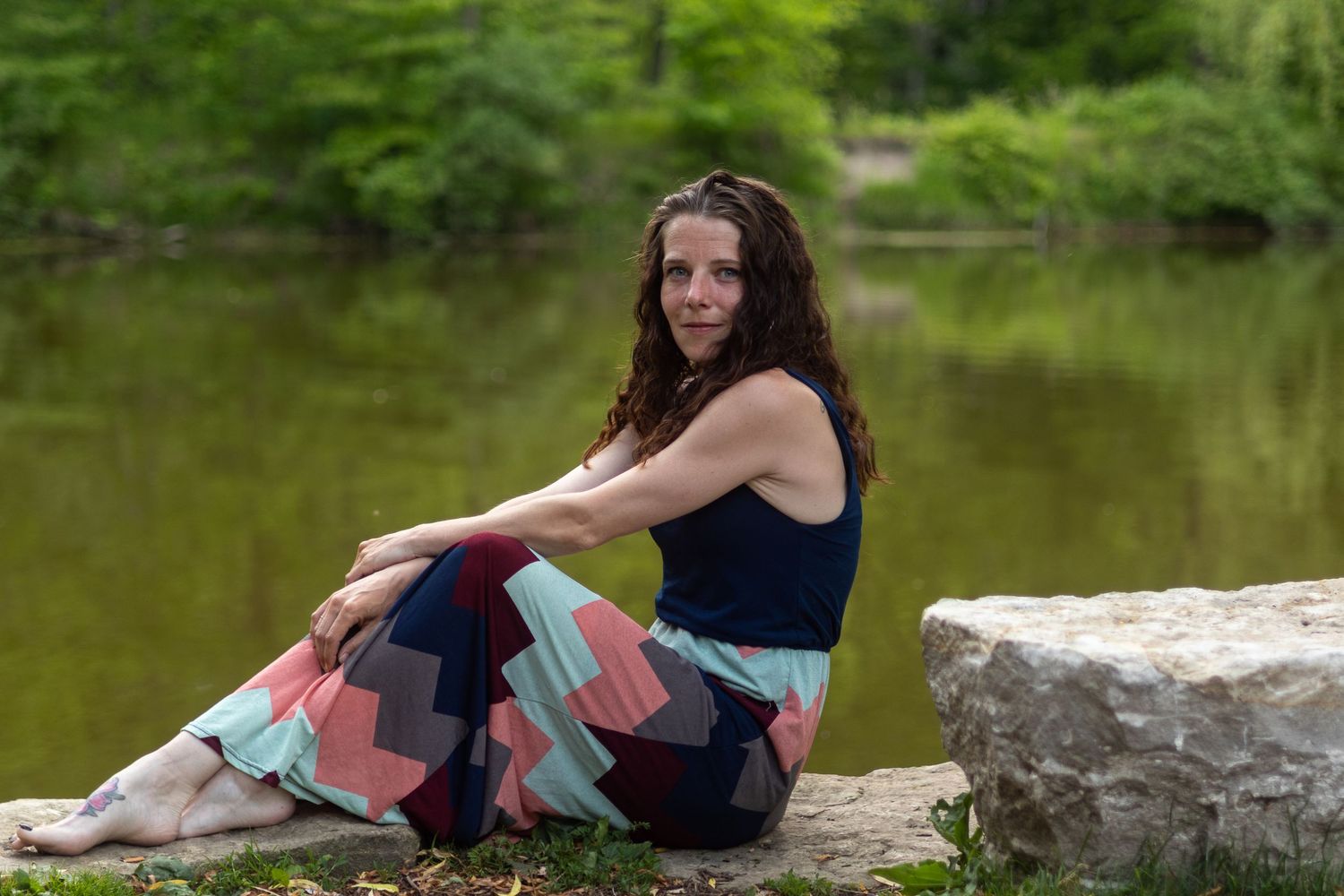 Woman in colorful patterned pants sits barefoot on rocks by a serene green lake surrounded by lush trees.