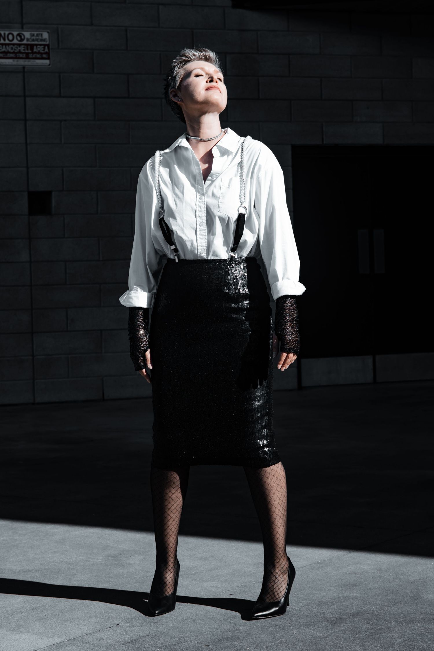 Woman in white blouse, black sequin pencil skirt, fishnet tights, and heels poses dramatically in sunlight against dark wall.
