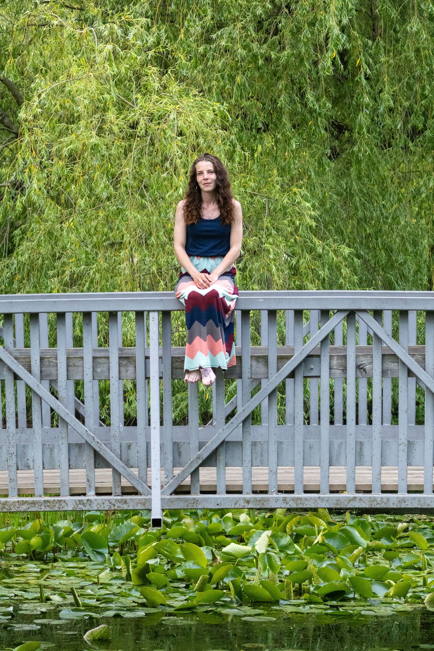 Young woman in colorful skirt sits on wooden bridge railing over lily pad pond with weeping willow backdrop.