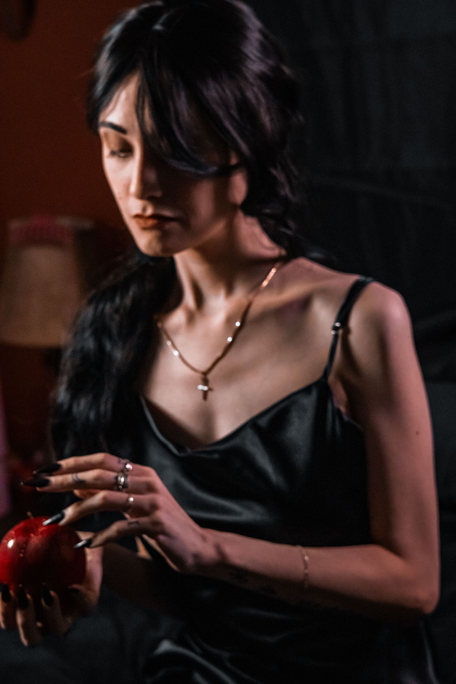 Woman in black satin dress holding red pomegranate, wearing gold necklace and rings in dramatic dark lighting.