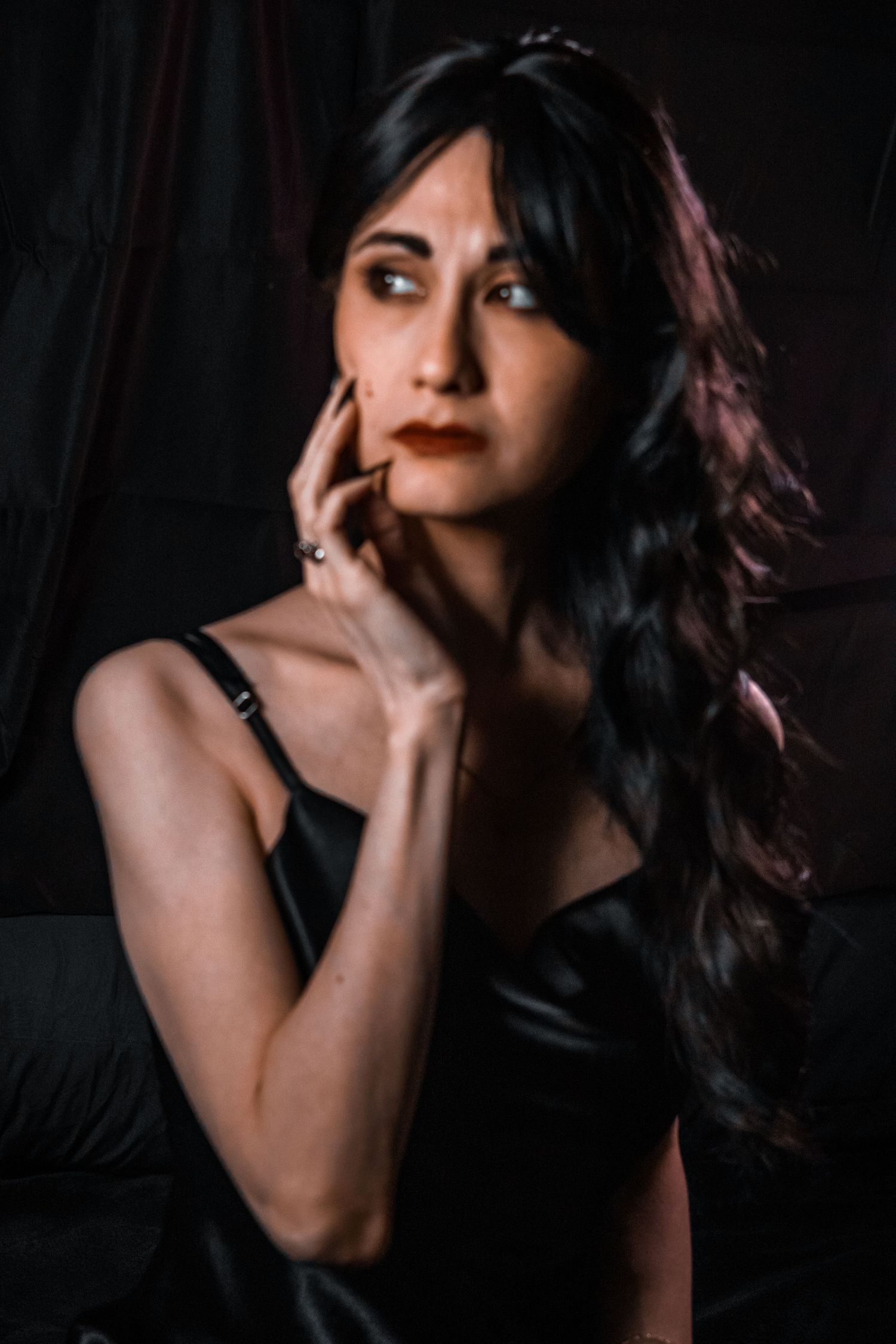 A woman with long dark wavy hair in a black satin dress poses thoughtfully with hand near face against dark background.