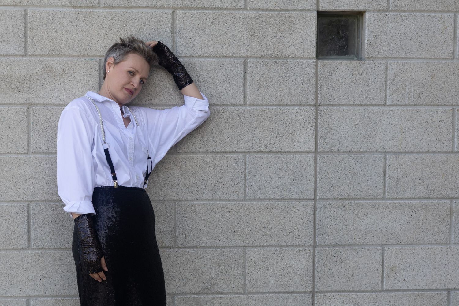 Person in white shirt, suspenders, and black gloves leaning against a concrete block wall.