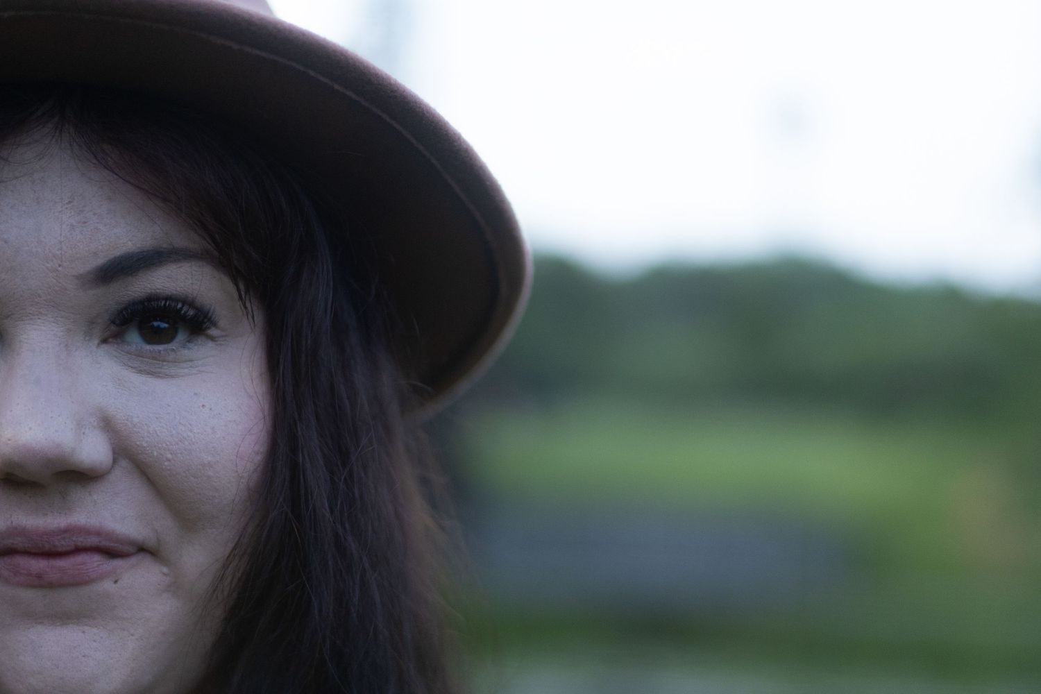 Close-up portrait of a young woman wearing a wide-brim brown hat with a blurred green landscape background.