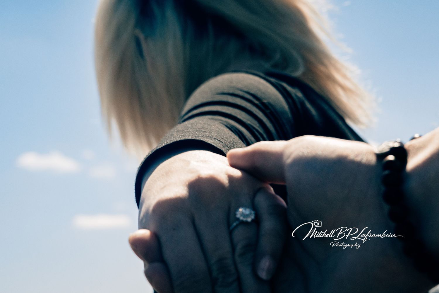 Close-up of couple holding hands showing an engagement ring against a blue sky background.