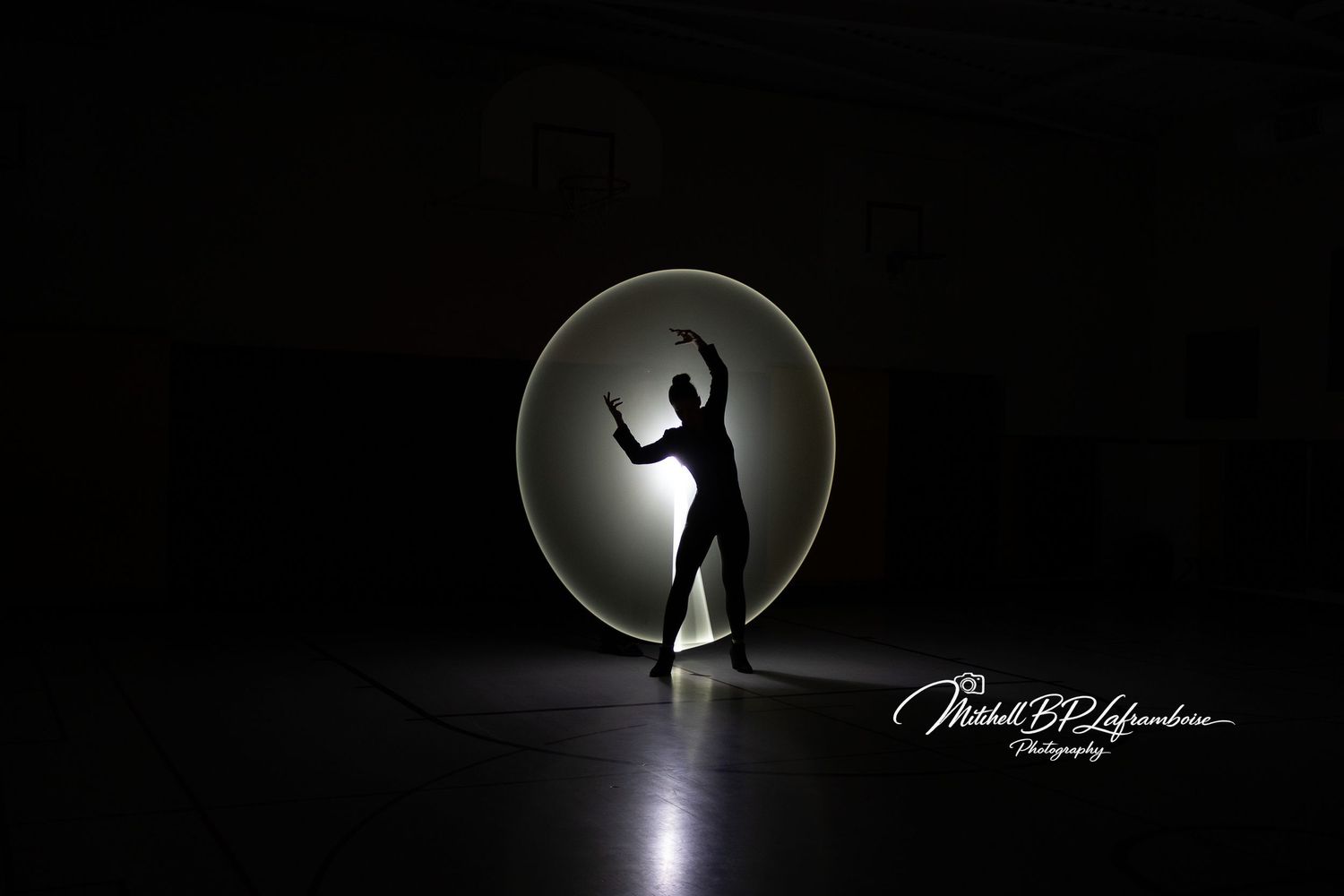 Silhouette of a dancer posed inside a glowing white circular light painting in dark gym.
