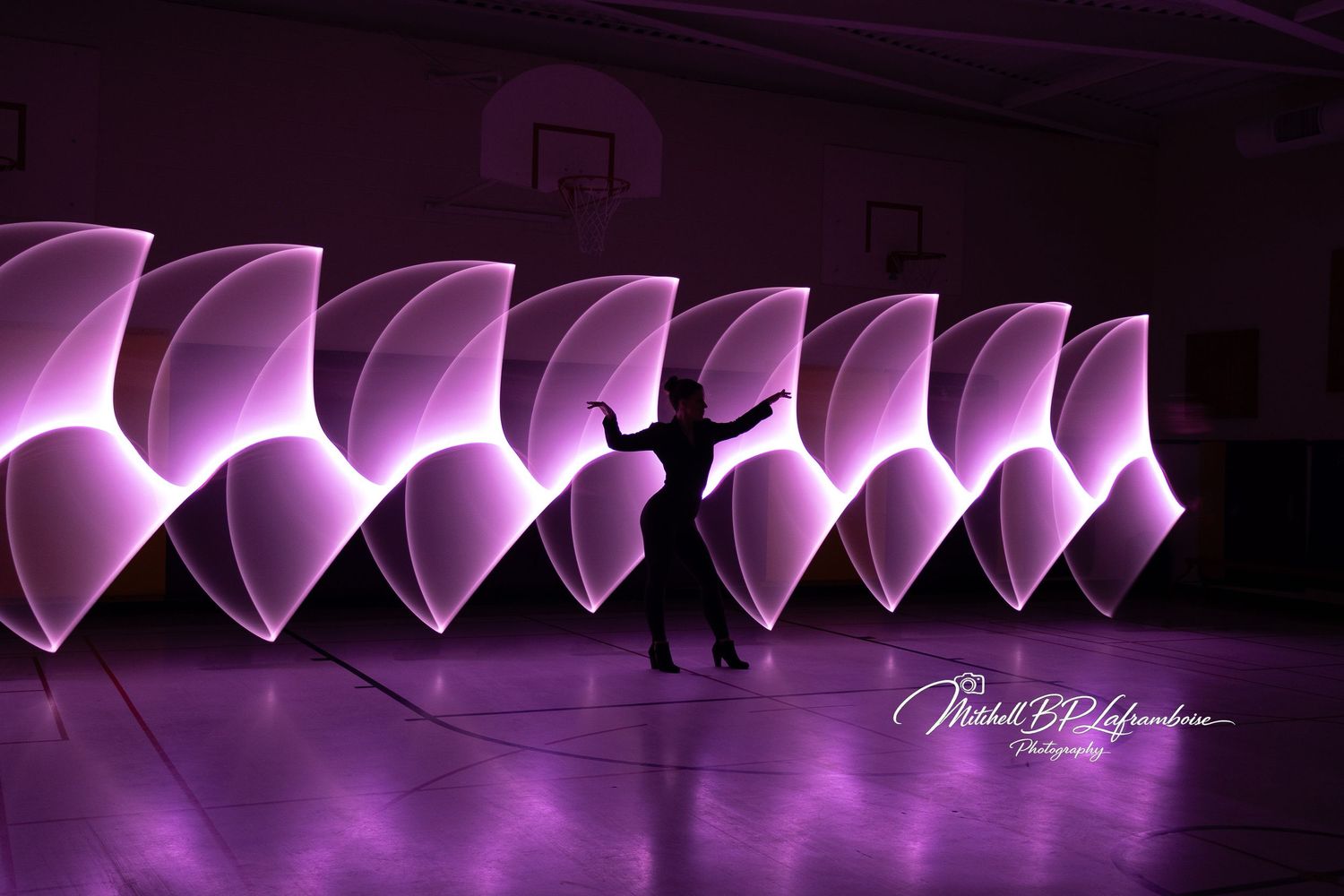 Dancer silhouette surrounded by repeating purple glowing petal light painting shapes in gymnasium.