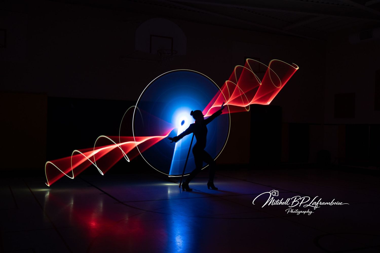 Silhouette of a person with red light painting spiral and diagonal streaks in dark gym.