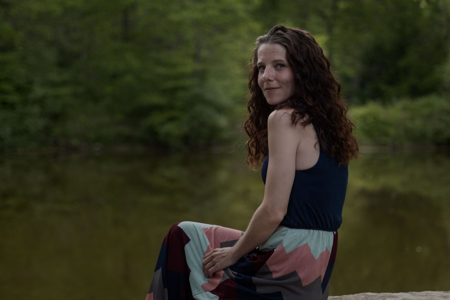 Woman with curly hair sitting on rocks by a serene forest pond, wearing a colorful maxi skirt and navy top.