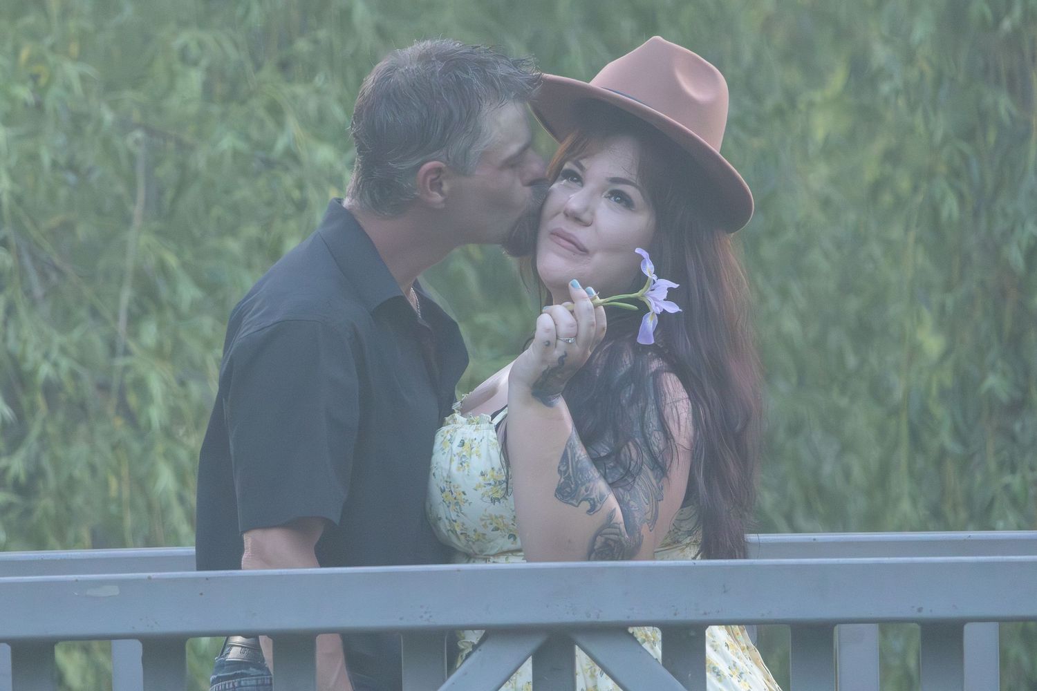 Romantic couple sharing a tender moment on a bridge, woman in floral dress and brown hat holds a flower.