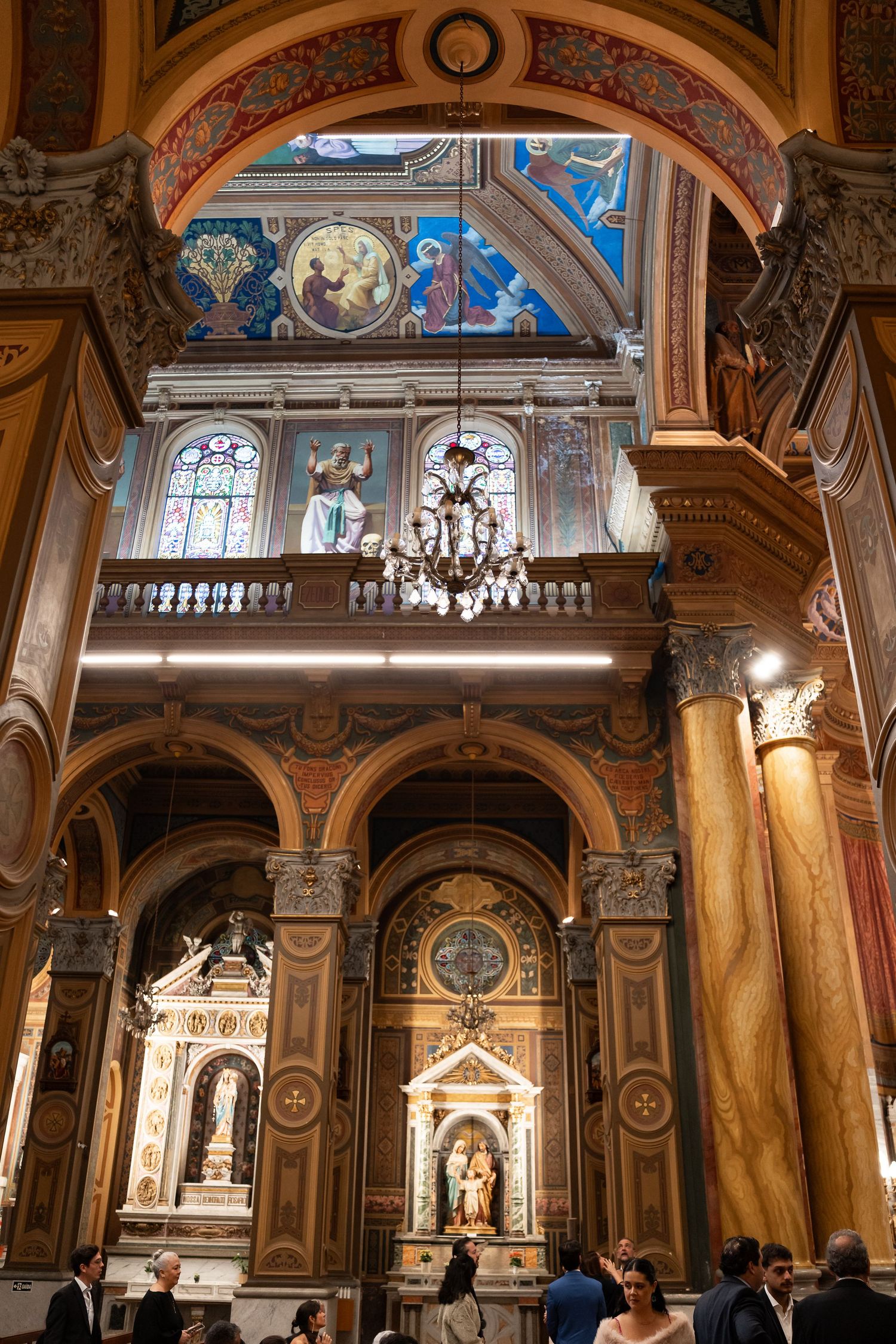 Ornate Catholic church interior with gilded altars, marble columns, colorful frescoes, stained glass windows, and a crystal chandelier.