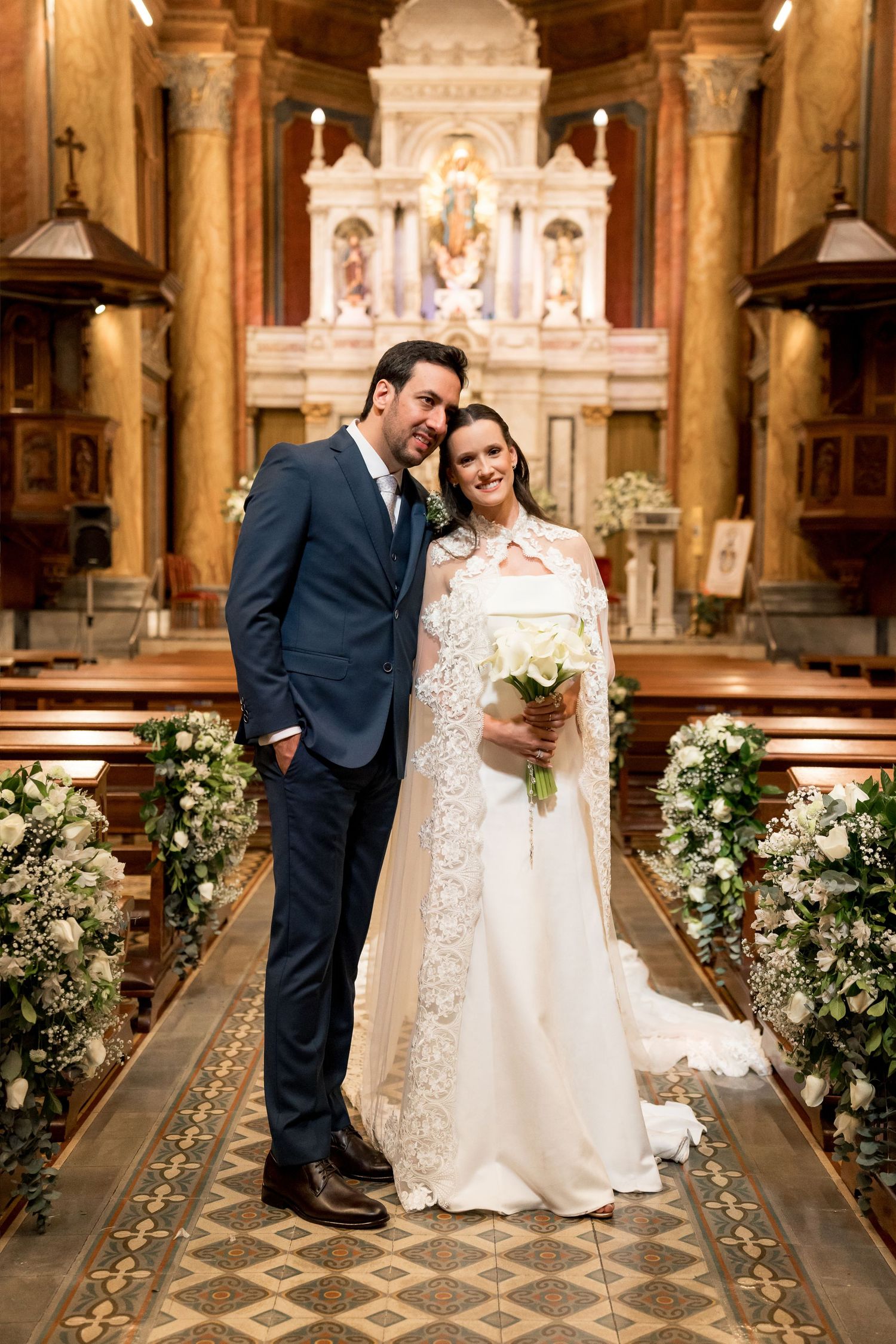 Bride and groom pose together at church altar, bride holding white bouquet in elegant lace wedding gown.