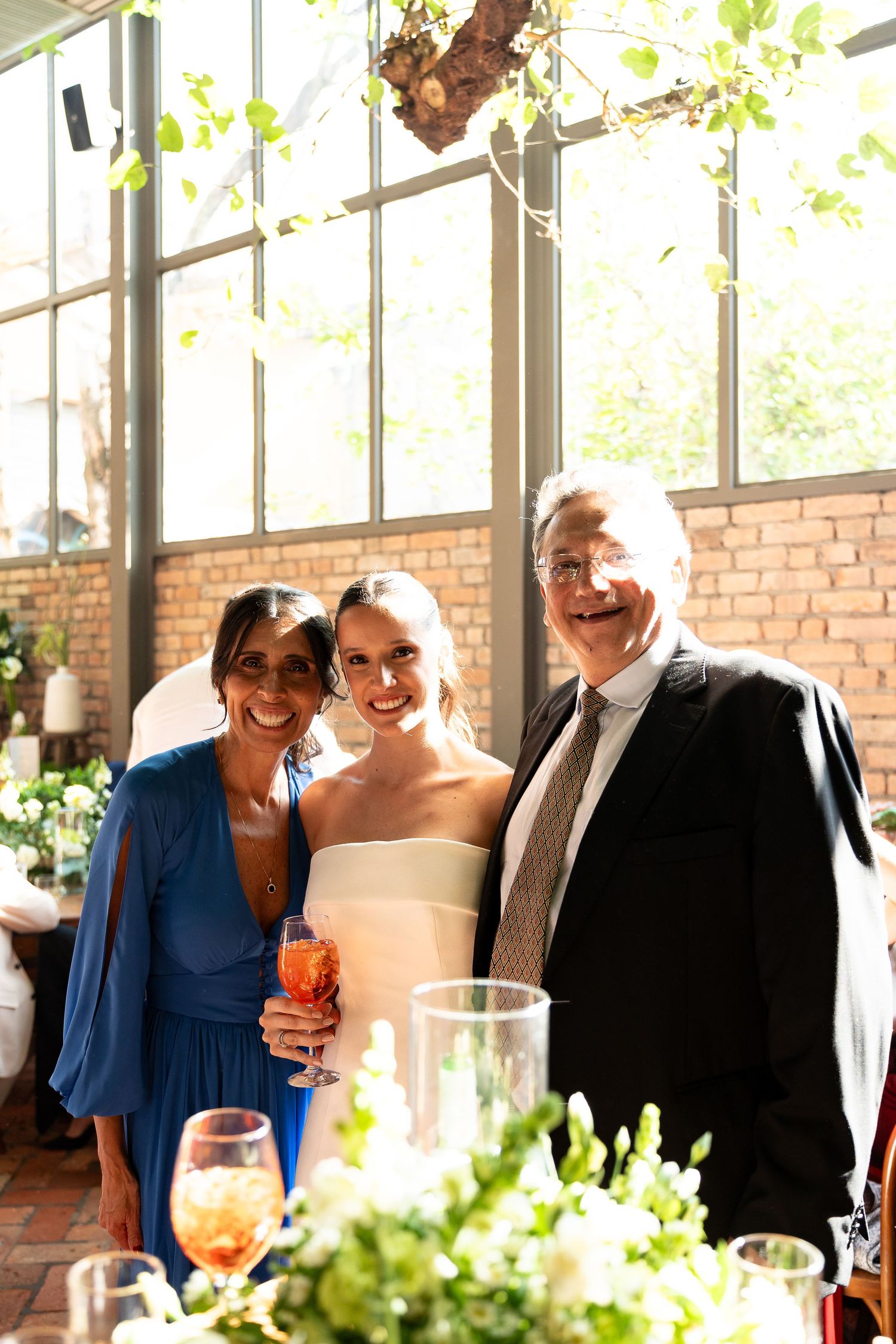 Three guests pose together at a wedding reception, with the bride in white holding a cocktail in a sunlit venue.