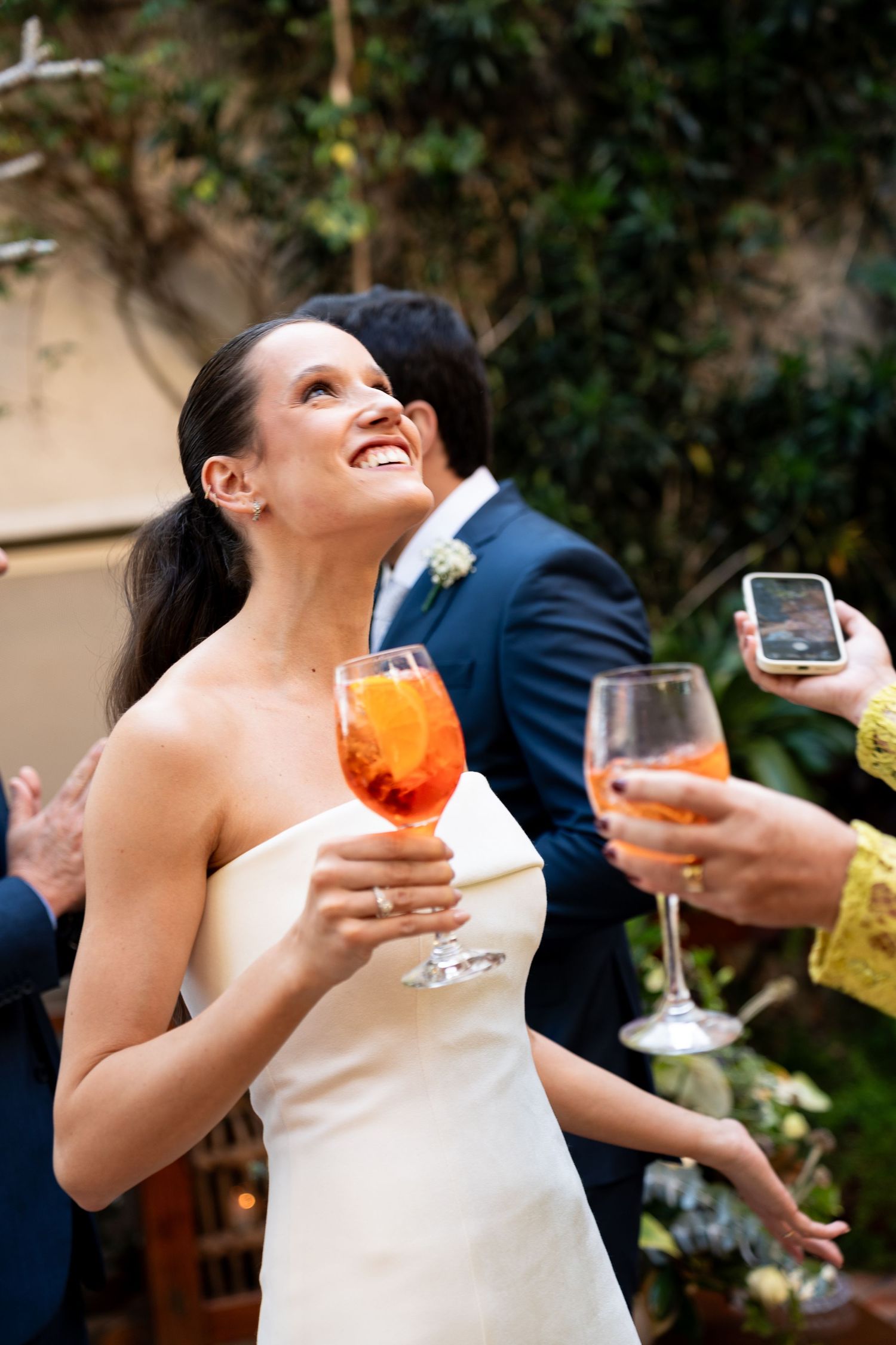 Smiling bride in white gown holding orange Aperol spritz cocktail at outdoor wedding reception.