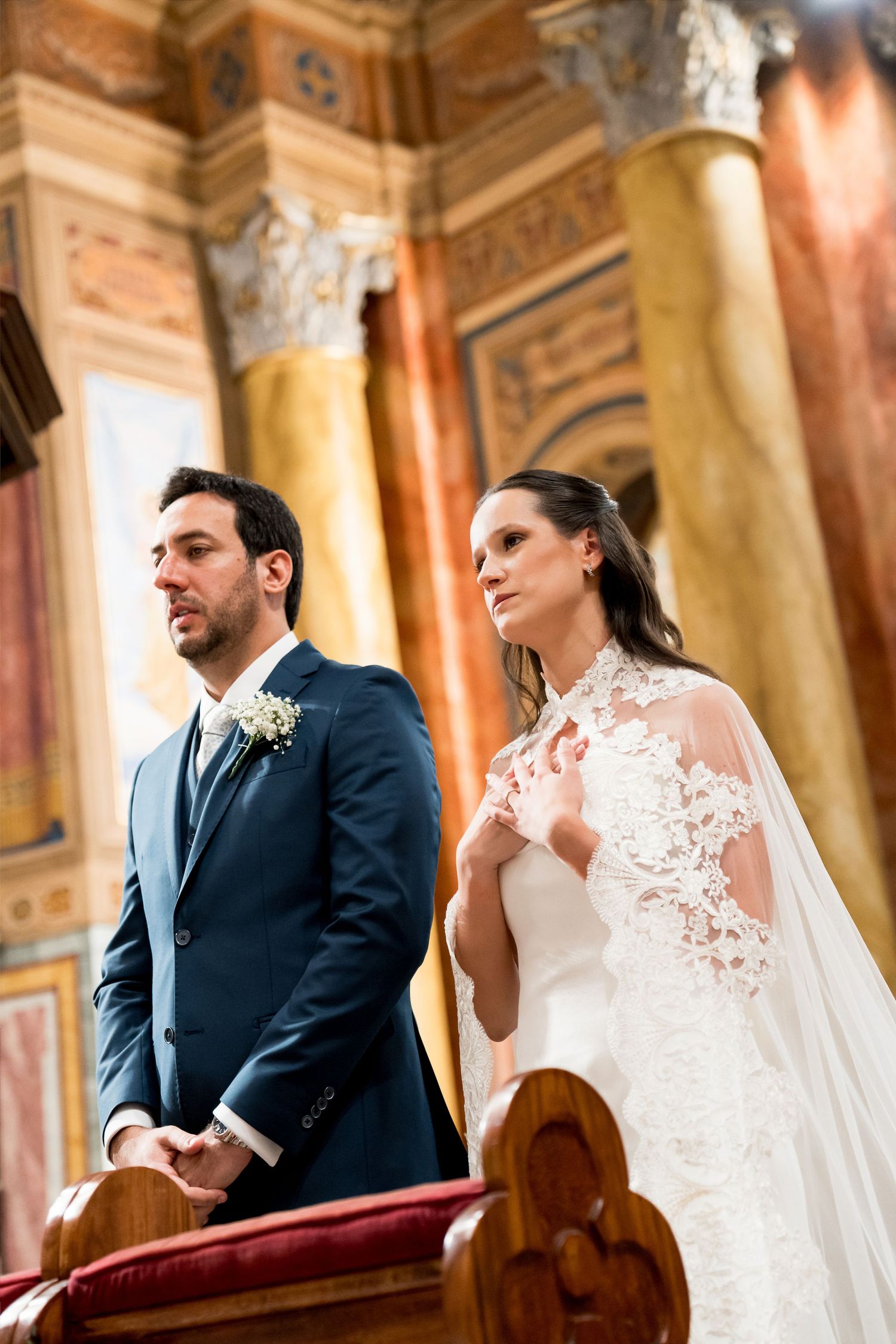 Bride in lace gown and groom in navy suit standing at church altar during wedding ceremony.