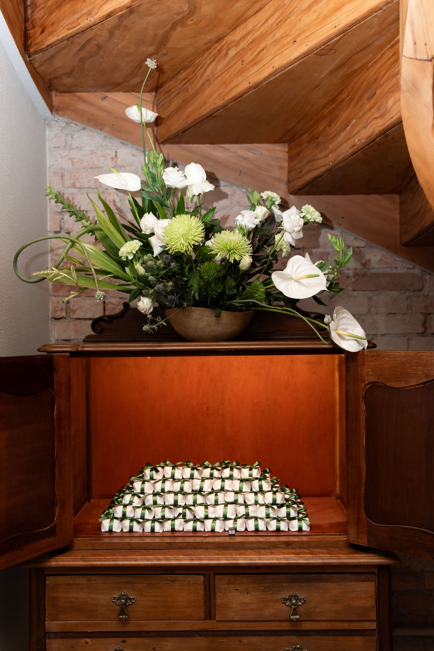 Wooden cabinet with floral arrangement of white anthuriums and green chrysanthemums atop, white roses inside, under wooden stairs.