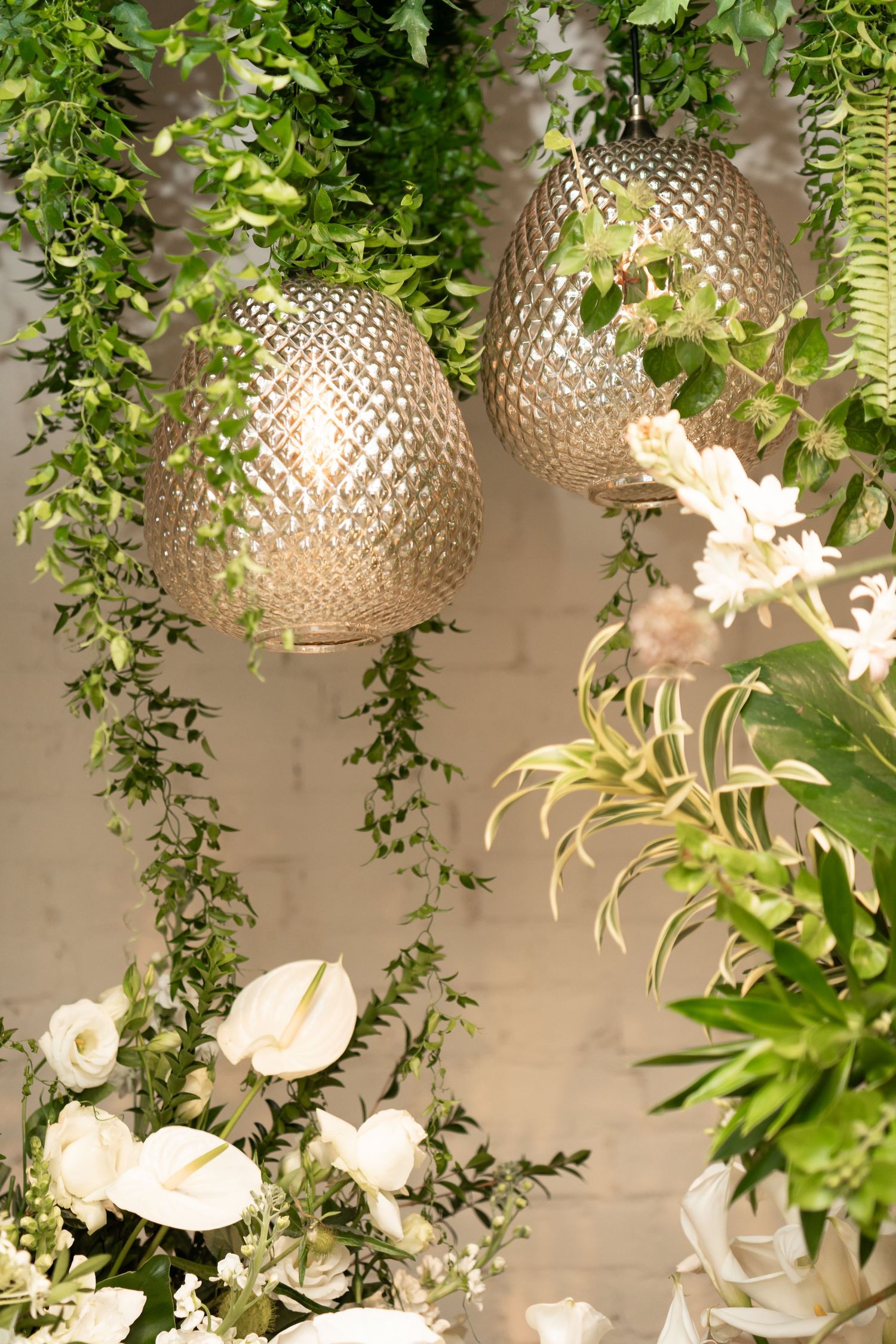 Two gold diamond-pattern pendant lights hang among lush green vines and white flowers in an elegant floral display.
