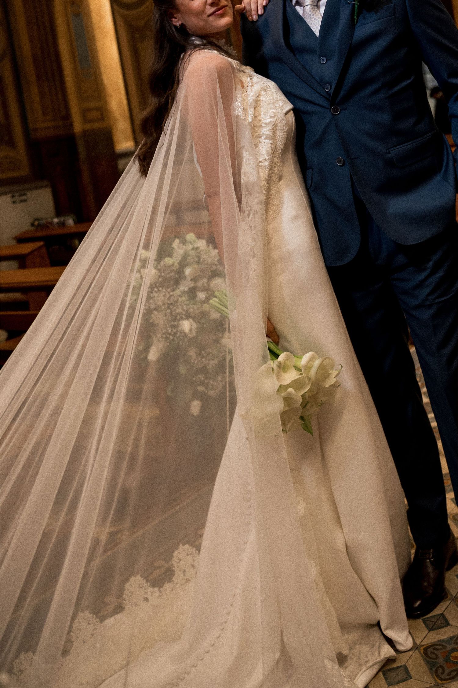 Bride in lace wedding gown with sheer veil holds white bouquet beside groom in navy suit at church ceremony.