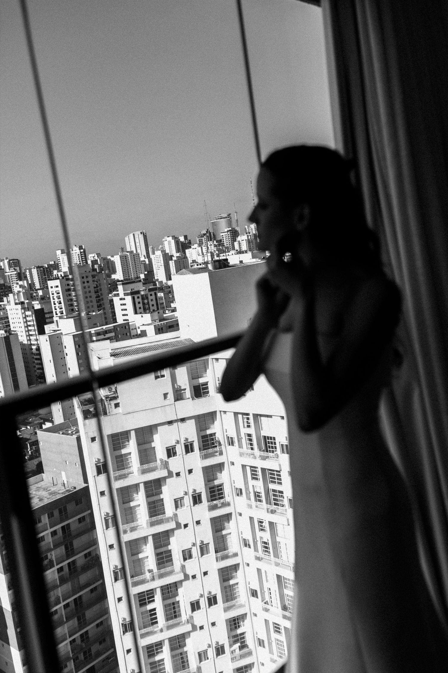 Black and white photo of a bride in wedding dress standing by a high-rise window overlooking a city skyline.