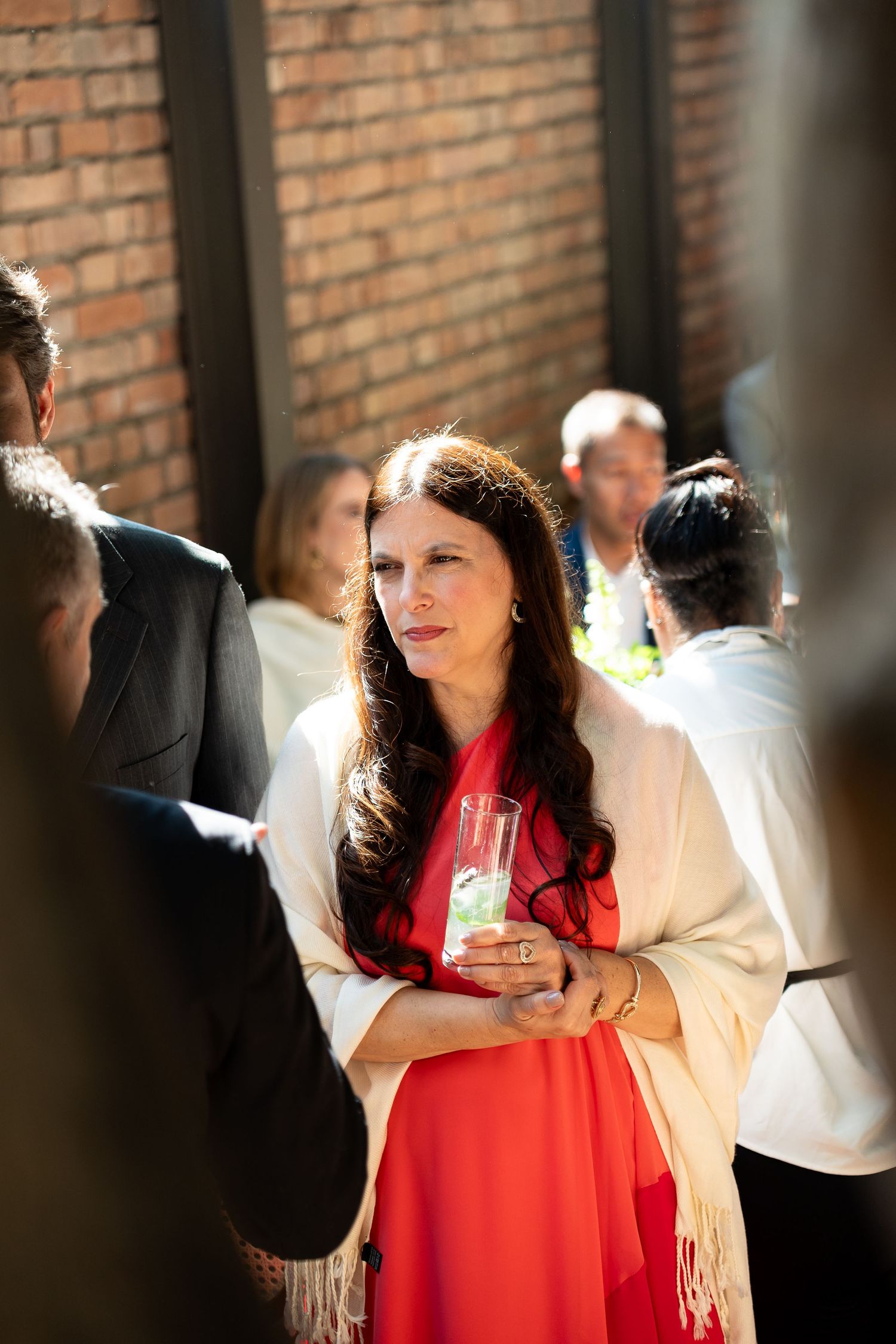 Woman in red dress and white shawl holding a drink at an outdoor social gathering near a brick wall.